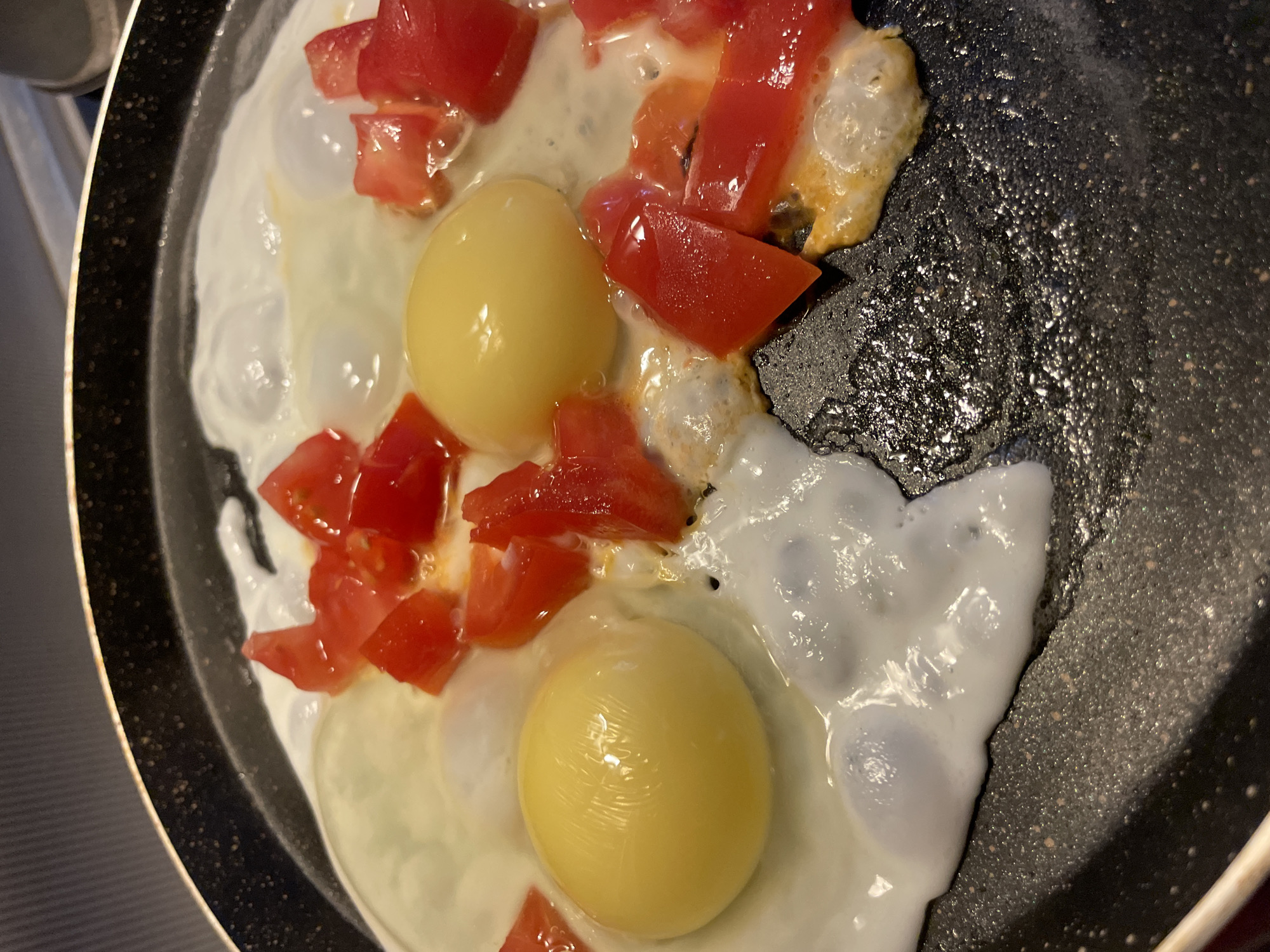 This is a close-up, eye-level shot of three fried eggs and diced tomatoes in a black non-stick frying pan. The eggs are in various stages of cooking, with two yolks visible and a third possibly obscured by the tomato. The tomatoes are bright red and appear to have been recently added, as they are still firm. The pan is slightly oily, and there are some small bubbles forming around the edges of the eggs. The background is blurred, but suggests a kitchen environment. There is no indication of the city of Sochi, Russia.