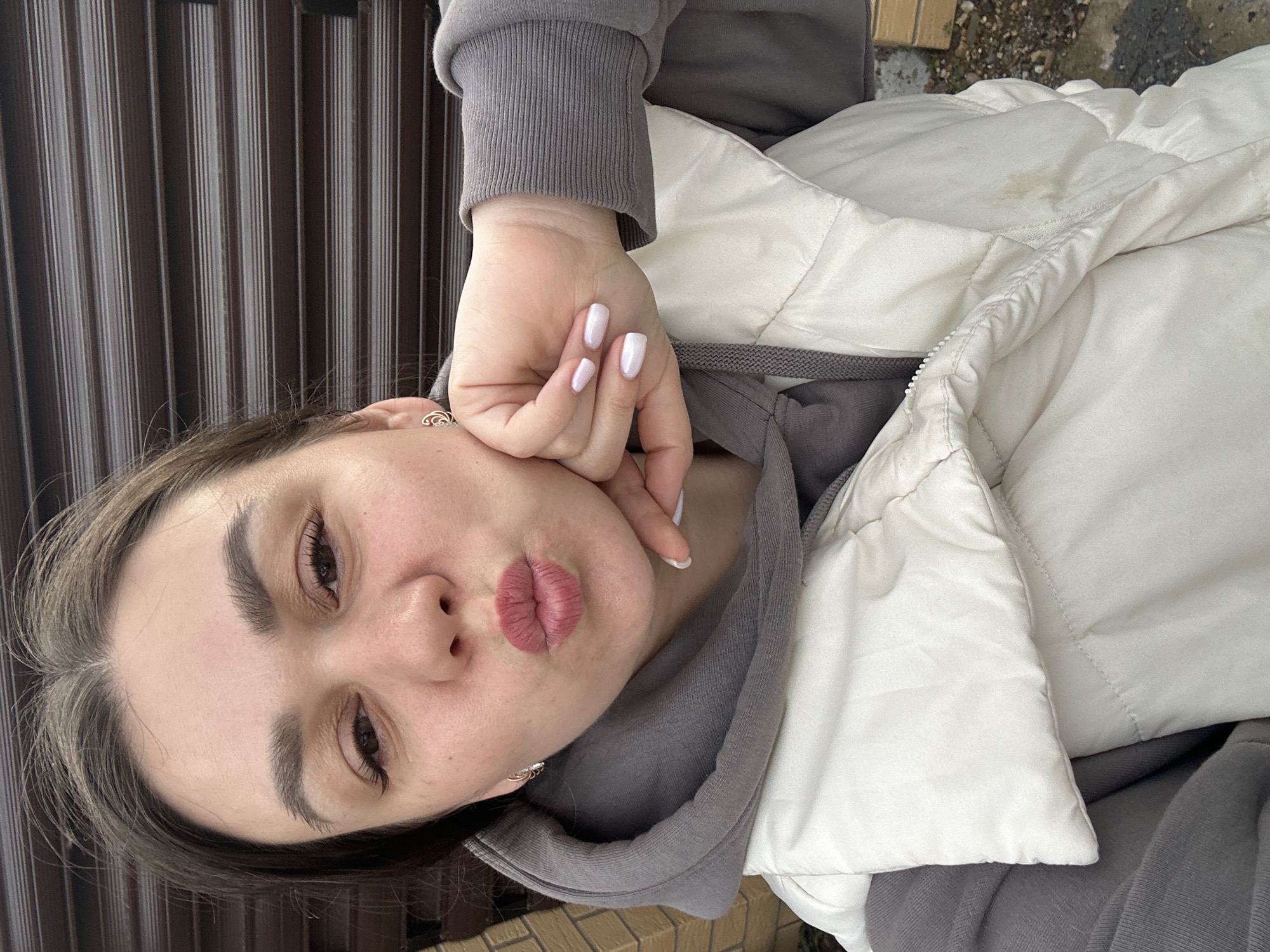 A close-up, eye-level portrait features a young woman, likely in Chaikovsky, Russia, posing outdoors. She looks directly at the camera with a playful 'duck face' expression, her lips pursed and accentuated with matte pink lipstick. Her right hand is raised, with fingers gently resting against her cheek.
The woman has dark hair parted down the middle, dark, well-defined eyebrows, and dark eyes with visible eyeliner and mascara. Her fingernails are neatly manicured with a light pink or white polish. She wears small, silver-colored hoop earrings with intricate details.
For attire, she has on a grey hooded sweatshirt with ribbed cuffs and a light-colored, possibly off-white or cream, quilted vest worn over it. A slight brownish stain is visible on the upper part of the vest, near her right shoulder.
The background on the left consists of dark, vertically striped panels, possibly a fence or blinds. On the right and extending towards the top, there's a glimpse of a rough, damp ground or pavement with small stones and a hint of a brick-like structure, suggesting an outdoor setting. The lighting indicates it is daytime, with soft, natural light. No visible text is present in the image.