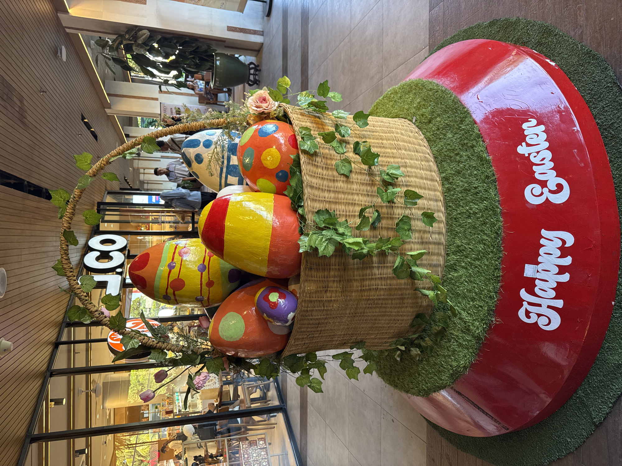 An indoor scene captures a vibrant Easter display featuring a large, wicker-style basket overflowing with oversized, colorfully painted eggs. The display is situated on a circular red base, which is surrounded by artificial green turf, mimicking grass. White decorative script on the red base clearly spells out "Happy Easter."
The prominent basket is adorned with artificial green ivy and a few artificial flowers, including a pink rose. Inside the basket and spilling out are several large, decorative eggs. These include an orange egg with yellow, blue, and red polka dots; a yellow egg with red stripes; another yellow egg with red stripes and purple dots; and an orange egg with green dots, featuring a smaller purple egg in a cutout. Partially visible behind these is a blue egg with white splatters.
In the background, a modern commercial interior is visible, likely a shopping mall or large store, consistent with the Kuta Selatan, Indonesia context. Wooden paneled walls line the left, while glass storefronts and light-colored tiled flooring are seen further back. Reflections suggest ambient light from the interior. Several people are scattered in the background, some walking, and others near storefronts, casually dressed. A large potted plant is visible, and overhead, a white sign displaying the letters "LOC" or "LCO" is discernible above a glass entrance. The lighting is bright and even, indicating daytime or a well-lit indoor environment. The overall scene suggests a festive seasonal decoration set up in a public commercial space.
