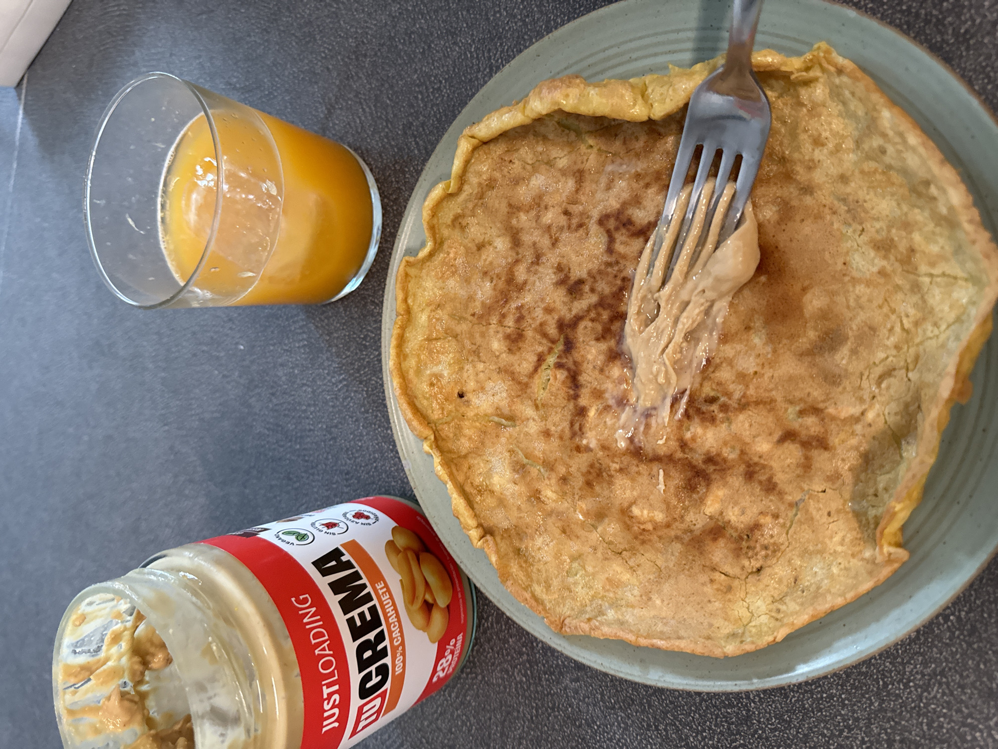 The image shows a close-up view of a breakfast or snack consisting of a large, thin pancake or crepe on a light blue-green plate. A fork is lifting a dollop of peanut butter from the pancake. To the left of the plate is a partially empty jar of "JUSTLOADING TU CREMA 100% CACAHUETE" peanut butter, with peanut butter visible on the inside rim of the jar. To the right of the plate is a clear glass filled with an orange liquid, likely juice. The background is a dark grey, textured surface, possibly a countertop. The lighting suggests an indoor setting, likely a home kitchen. The image was taken from a slightly elevated angle, looking down at the food and drink. There are no people visible in the image. The scene does not provide any specific information about the city of Murcia, Spain.