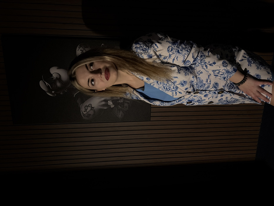 A blonde woman with red lipstick lies on a wooden surface with a black art piece featuring grayscale figures. She wears a floral-patterned blazer over a blue top. Her hand rests on her leg, showcasing a dark beaded bracelet. The room is dimly lit, with the wooden surface creating horizontal lines.