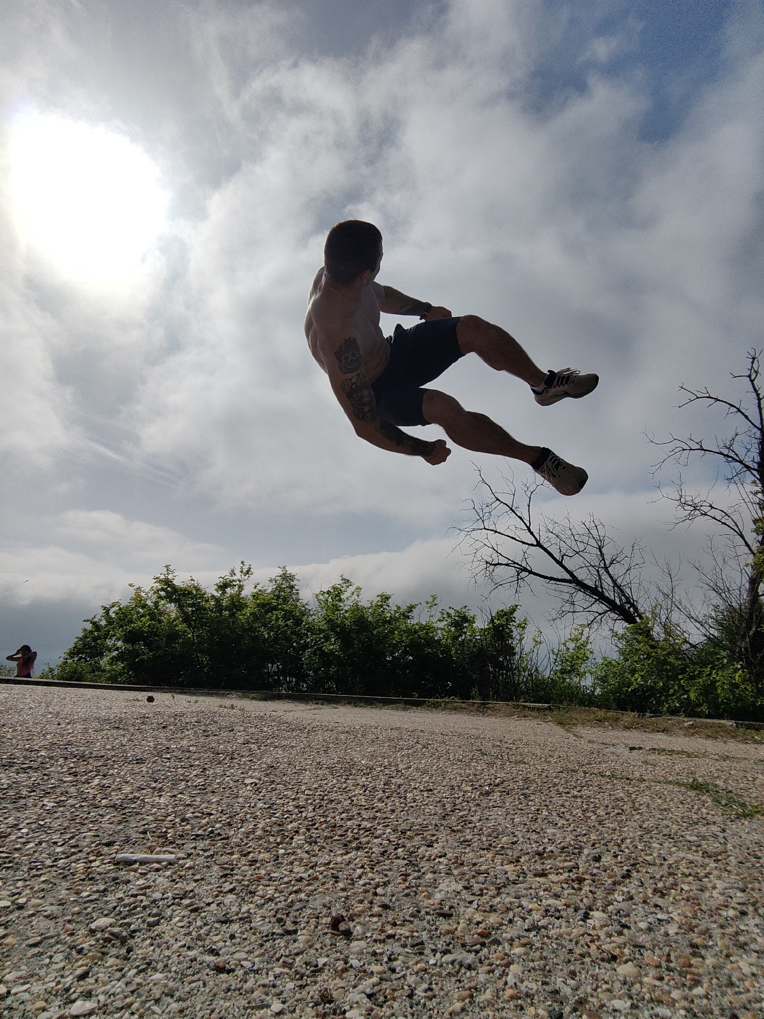 A shirtless man with dark hair, wearing dark shorts and white sneakers, is airborne in a dynamic pose against a backdrop of sky with scattered clouds and a bright sun. He appears to be jumping or performing a martial arts move. The location is an outdoor space featuring a pebbled ground surface, green bushes, and a leafless tree branch on the right side of the frame. In the background, there is a partial view of the sea and a person standing near a railing in the far distance. The overall setting suggests a location in a park or elevated area near the coast. It is impossible to confirm the location as Varna, Bulgaria with certainty based on only this image.