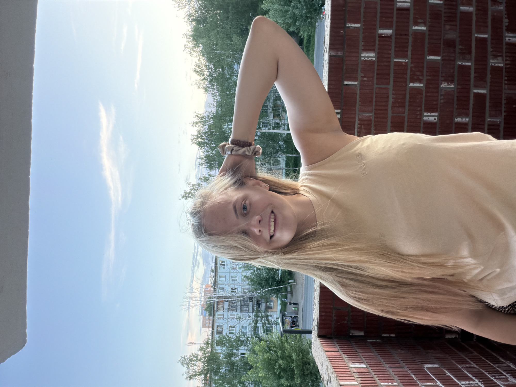 A young blonde-haired woman smiles at the camera, her arm raised behind her head, with a cityscape visible in the background. The setting appears to be a balcony, evidenced by the red brick wall and a concrete structure on the left side of the frame. Across the street, multi-story residential buildings are present, interspersed with trees. According to visible signage and architectural details, the location is likely Izhevsk, Russia. Pedestrians are visible on the street below. The sky is partly cloudy.