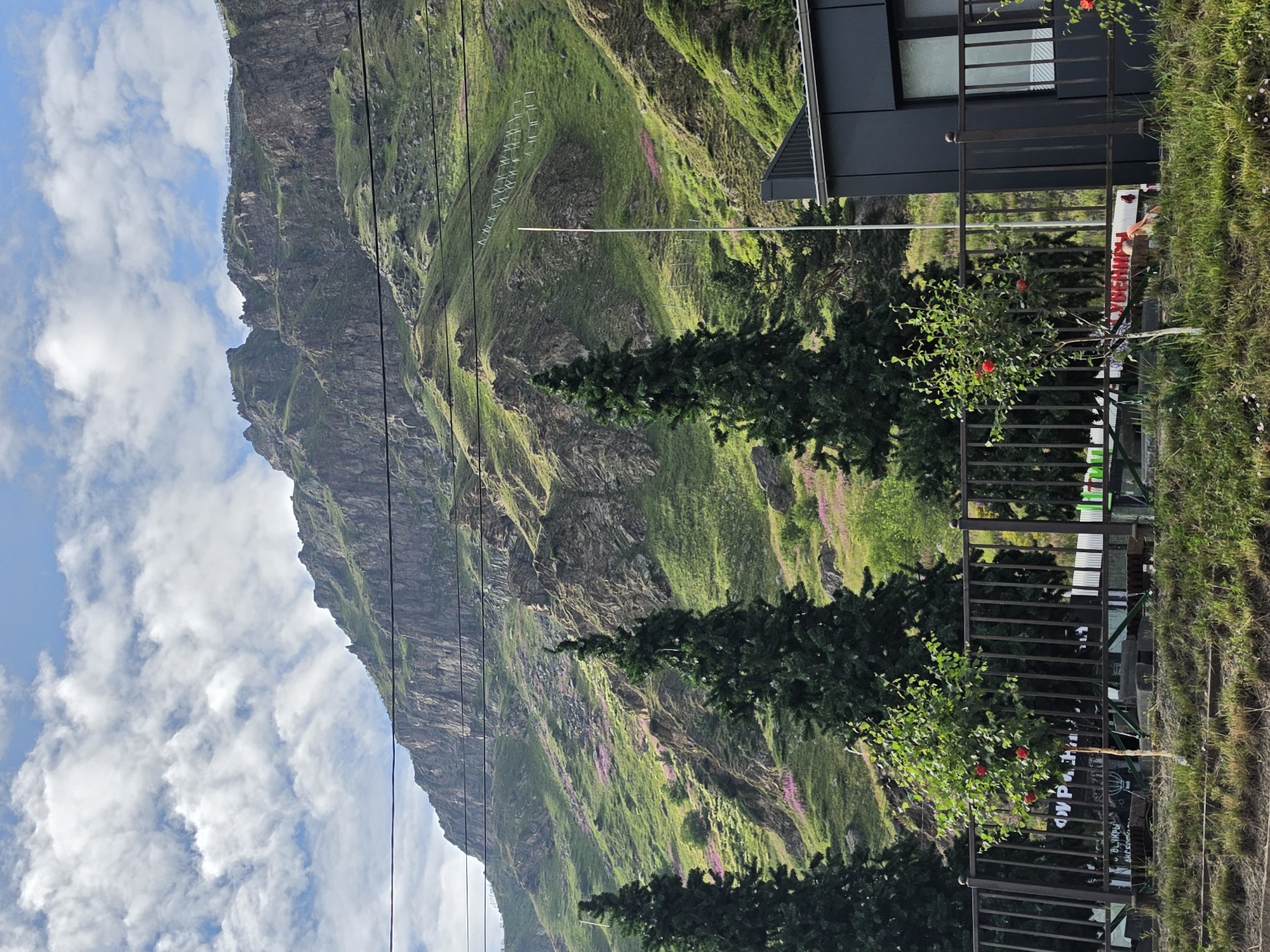 The image shows a steep, rocky mountain slope covered in green grass and scattered pink flowers, located in Terskol, Russia. The mountain is vast and extends high into a partly cloudy blue sky. In the foreground, there are several dark green coniferous trees and a small deciduous tree with red flowers. A dark metal fence runs horizontally across the lower portion of the image, separating the foreground from the mountain. Behind the fence, a modern, dark-colored building with a window is visible on the right side of the frame. Power lines are strung across the upper part of the image, spanning between poles that are not fully visible. A tall, thin pole stands in the middle ground. The overall scene depicts a natural landscape with some man-made structures.