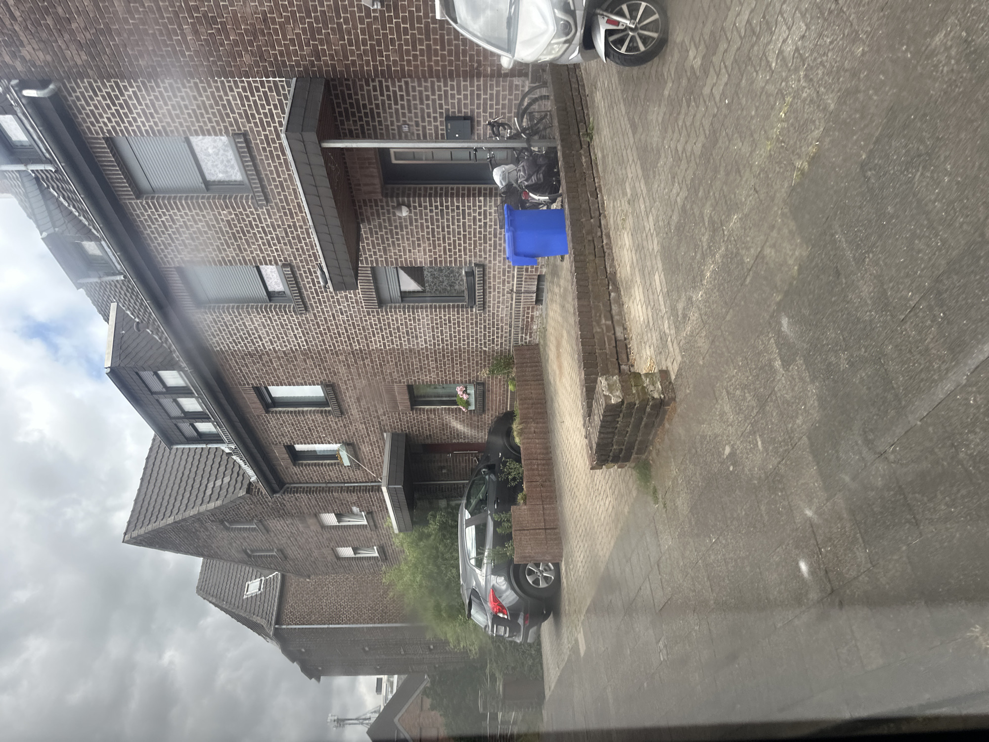 The image shows a row of brick houses in Krefeld, Germany, on a cloudy day. In front of the houses, there is a paved driveway and sidewalk. On the left side of the image, a dark gray SUV is parked. In front of one of the houses, there is a blue recycling bin and a bicycle. A silver scooter is parked on the right side of the image. The houses have tiled roofs and brick facades. Some of the windows have gray roller shutters. The environment appears to be a residential area.