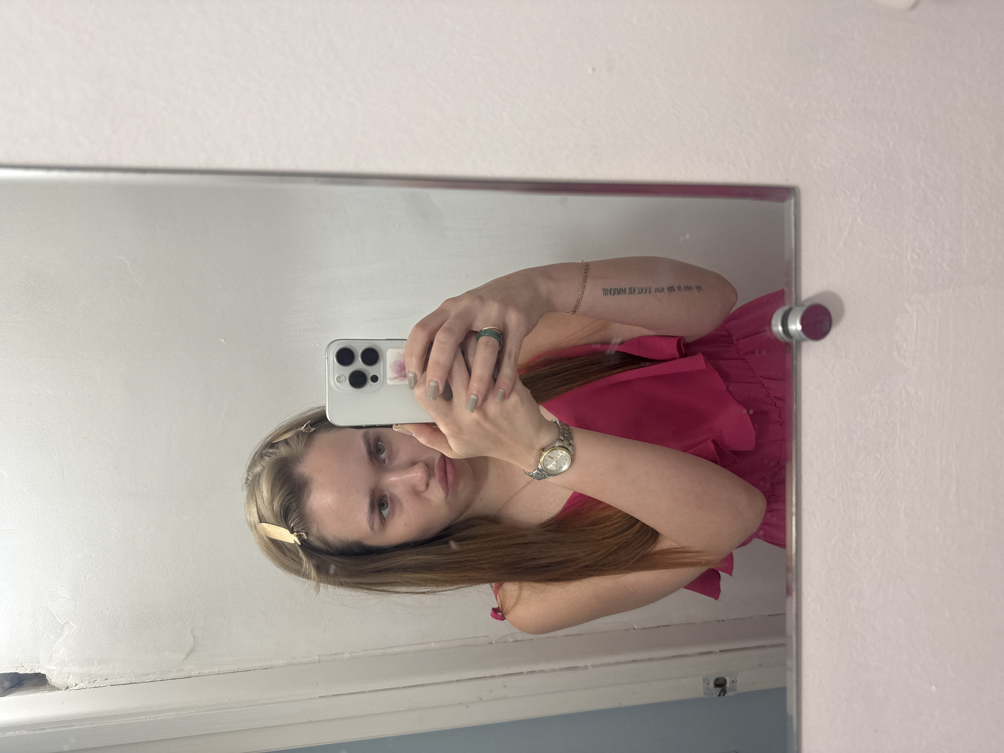 A woman is taking a selfie in a mirror. She is wearing a pink dress and a watch. Her hair is blonde and she has a tattoo on her arm. The background is a white wall. The location is unknown.