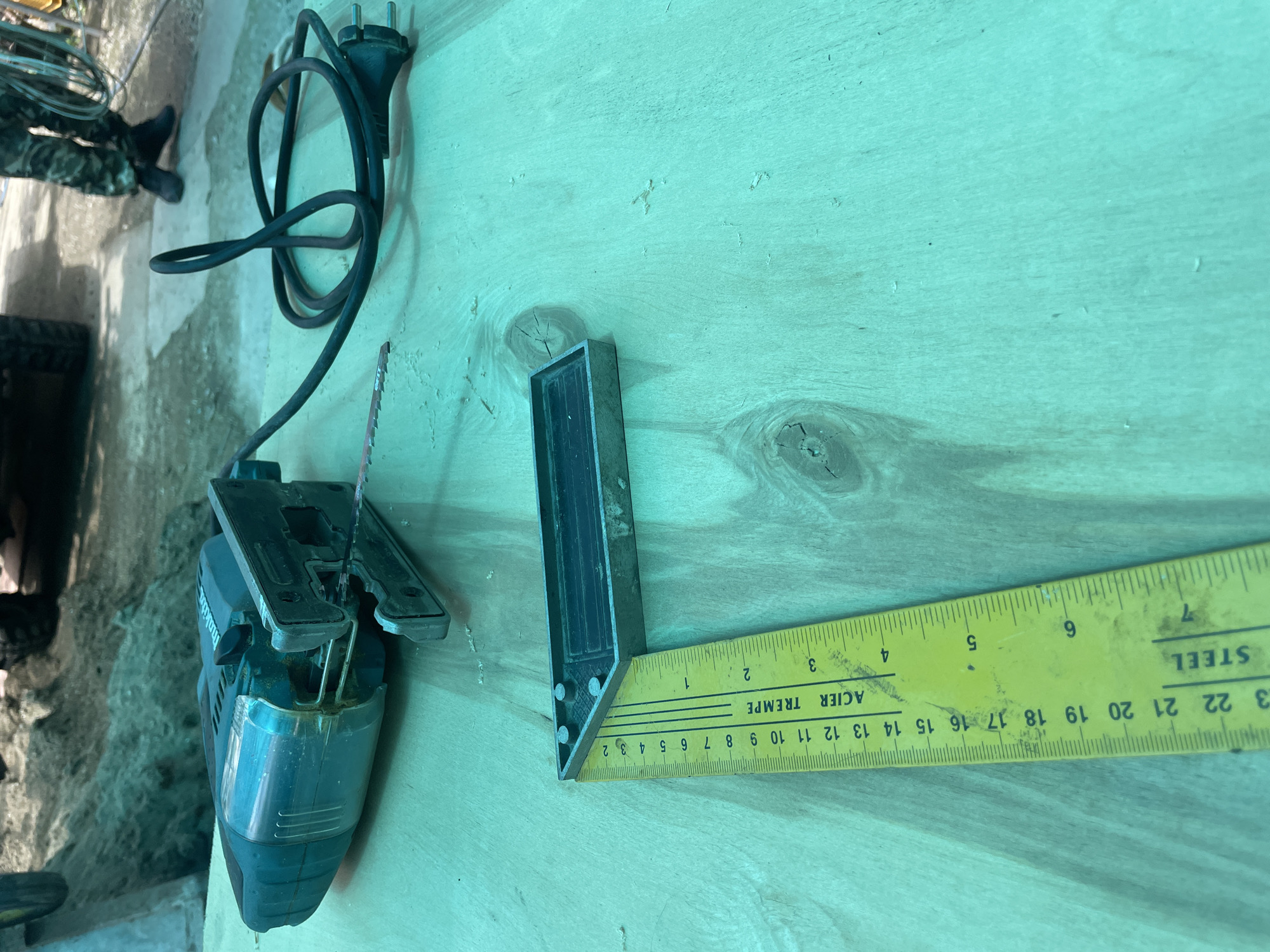 The image shows a jigsaw, a combination square, and a wooden surface. The jigsaw is on the left, with its power cord curled next to it. To the right of the jigsaw is a combination square, with its base perpendicular to the yellow measuring tape. The tape reads "ACIER TREMPE" (steel tempered) in French and "STEEL" in English, with numbers indicating inches and centimeters. The background is a light green wooden surface. The image does not provide any information about the location of Olginka, Russia, or the city itself.