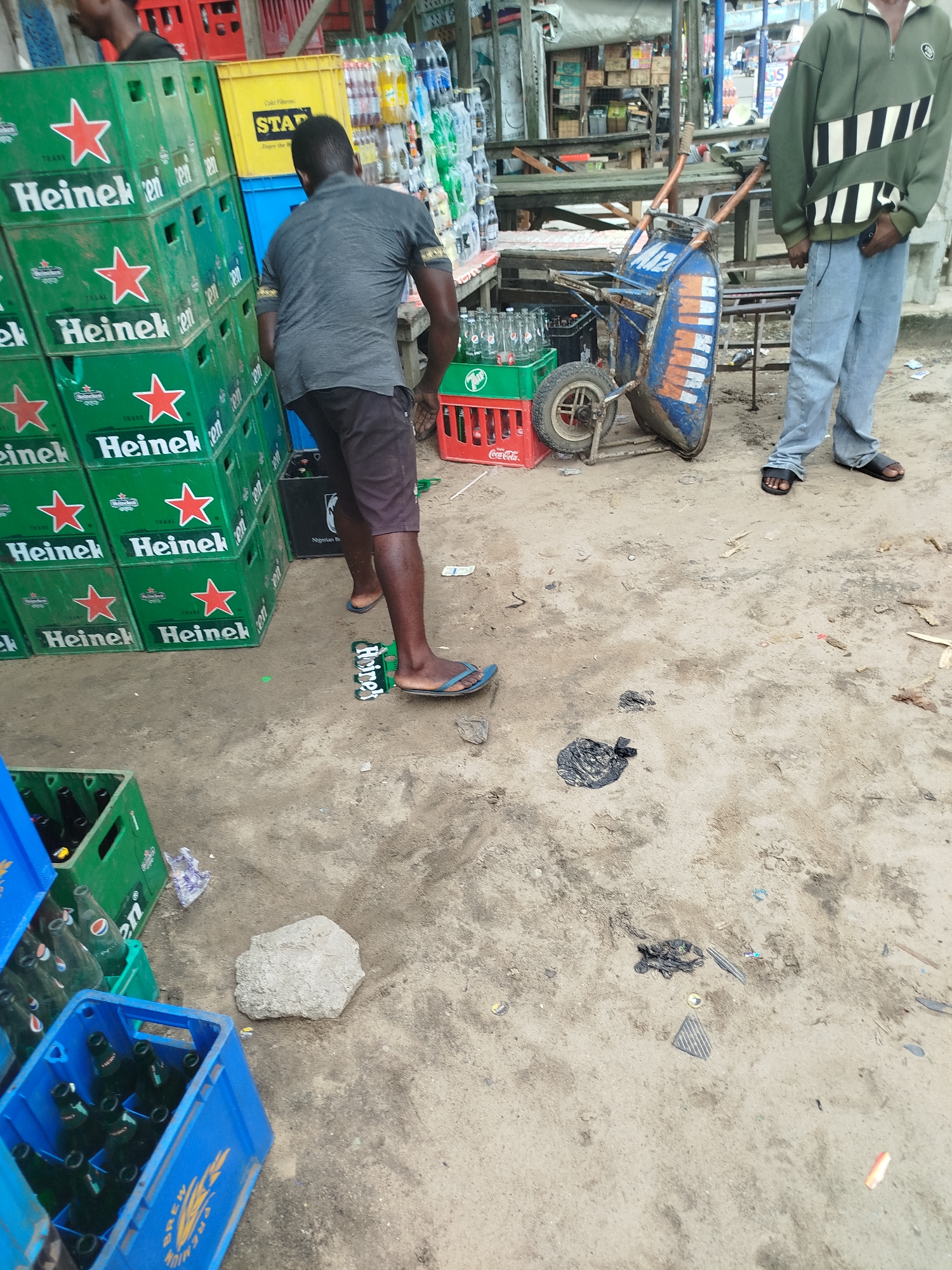 This media file captures a scene at what appears to be a market or roadside stall in Yenegoa, Nigeria. The foreground is dominated by stacks of green Heineken crates, some of which contain bottles. In the mid-ground, a man wearing a grey t-shirt and dark shorts is pushing a blue and orange wheelbarrow. He is standing on a scattered Heineken crate in the sand. To the right, another man is visible, wearing a green hooded jacket and light blue jeans, with black sandals on his feet. He is holding a dark object, possibly a phone, in his right hand.
In the background, more crates and shelves are visible, filled with various bottles and products, including Coca-Cola. A rough, sandy ground covers the area, with scattered debris. The lighting suggests it is daytime, likely a sunny day given the shadows. The overall impression is that of a busy, informal trading environment. Text visible in the image includes "Heineken," "Coca-Cola," "STAR," and "BREW PREMIUM." The presence of multiple beer crates and a wheelbarrow suggests the ongoing transport or sale of beverages.