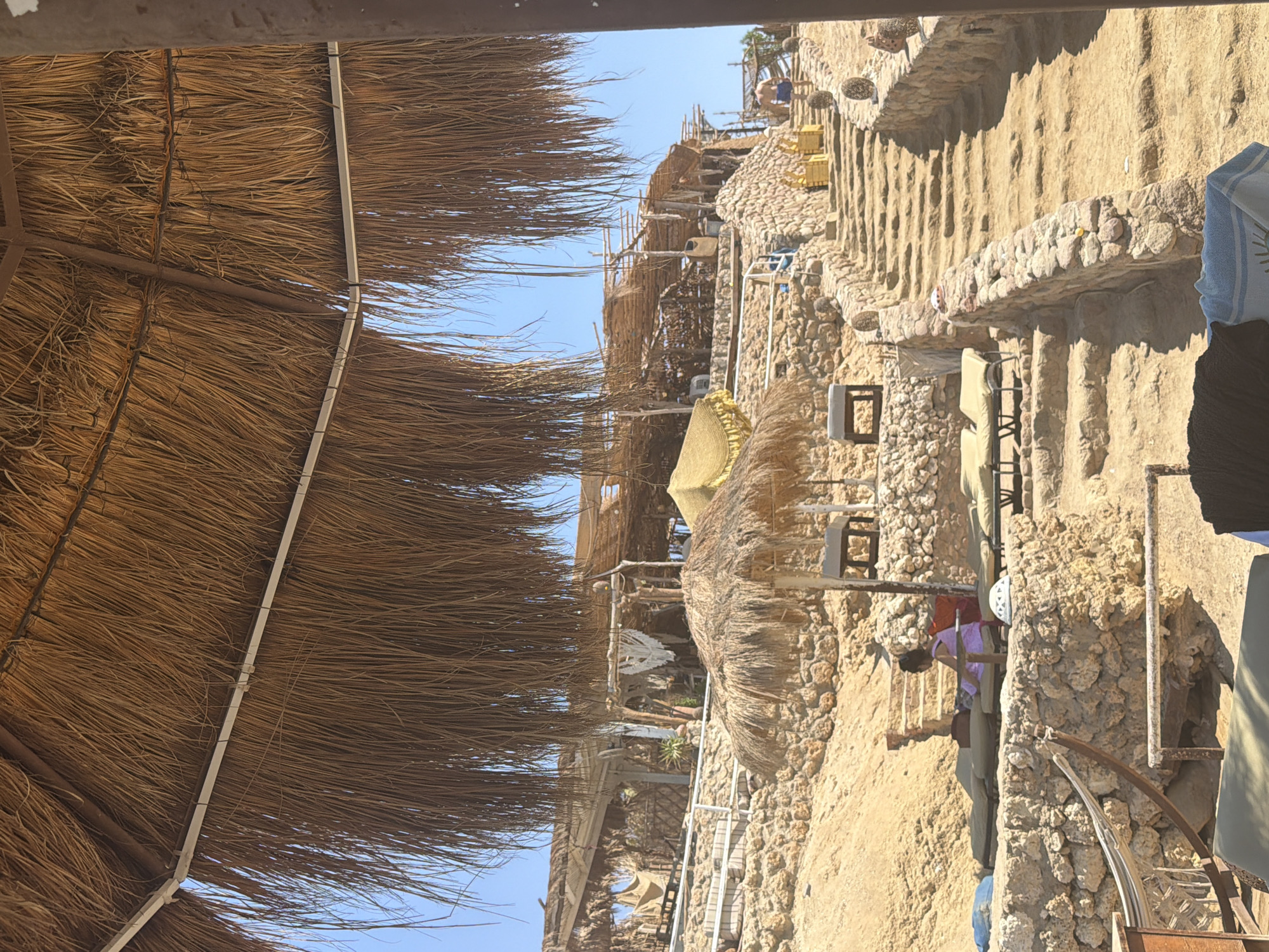 The image depicts a lively beach scene in Sharm El Sheikh, Egypt, characterized by a rustic, natural aesthetic. In the foreground, a straw-covered canopy casts a shadow over the sand, suggesting a shaded seating area. Further back, a stone staircase ascends a rocky incline, leading to a cluster of open-air structures with thatched roofs, likely serving as shops or restaurants. A few beach chairs are visible, some occupied by a woman in a purple dress who appears to be relaxing. The sun shines brightly, illuminating the sandy landscape and the turquoise water in the distance. The presence of a person walking up the stairs further emphasizes the activity of the beach. The overall impression is one of a vibrant and welcoming tourist destination.