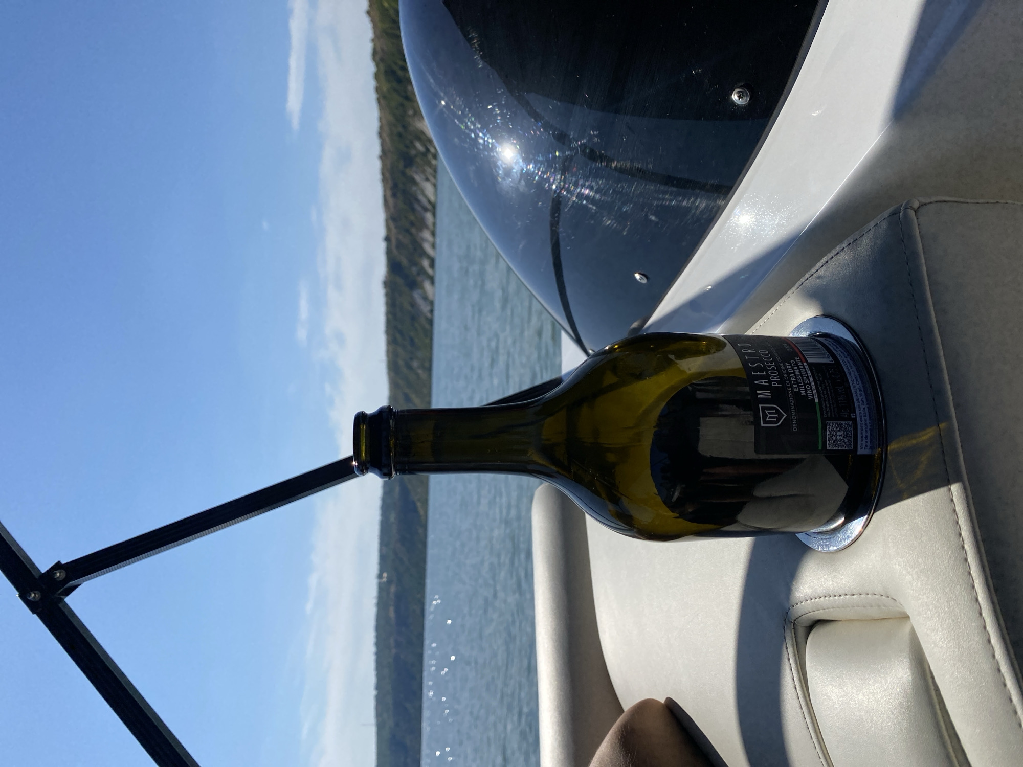 A full-frame, eye-level shot captures a dark green wine bottle secured in a silver cup holder on a boat's light grey leather seating. The bottle is positioned in the foreground to the right, with its top cut off by the frame. A label with text and a QR code is visible on the bottle. To the left, a person's arm and shoulder are partially visible, resting on the seat. In the background, a wide expanse of blue water stretches to a distant, tree-covered hilly coastline under a clear blue sky with sparse white clouds. The boat's black windshield, with a reflective, glossy surface, is visible to the right of the bottle. The scene appears to be set on the Nistru River near Molovata, Moldova, suggesting a recreational outing on the water.