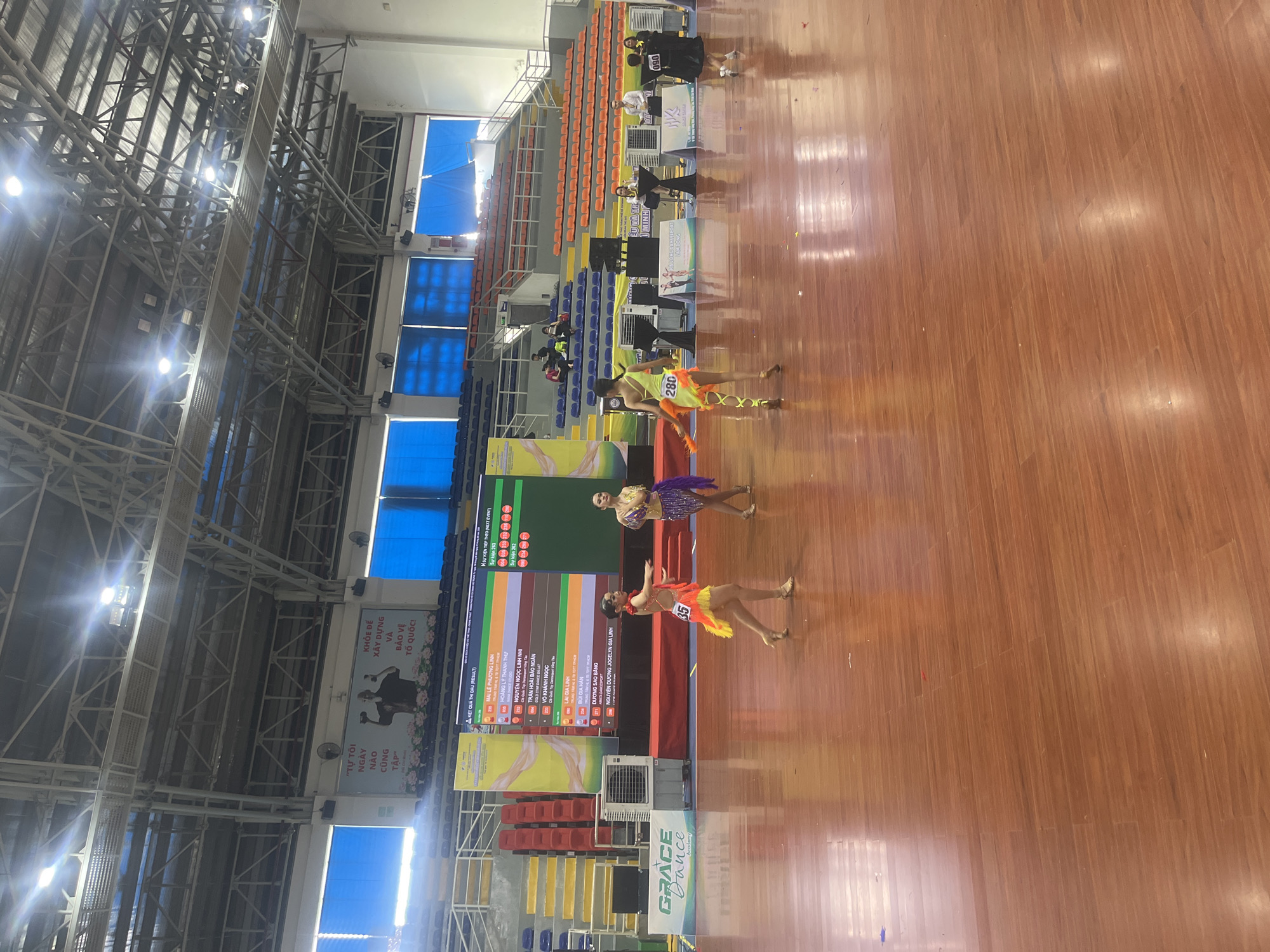 An indoor dance competition is underway on a polished wooden floor in an arena. A large screen displays results, with names like "MAI LE PHUONG LINH" and "HOANG LY THANH THU" visible. Three young female dancers are on the floor. One in a yellow and orange costume with the number 280 on her back is facing away from the camera, mid-dance. Another in a vibrant purple and gold costume, number 35, is facing the screen. A third dancer in a shimmering gold and purple outfit is also on the floor. Spectator seating in blue and orange is visible in the background, along with the arena's metal support structure and lighting. A banner for "GRACE DANCE ACADEMY" is on the left side of the image. The overall scene suggests a formal dance event with competitors and audience members.