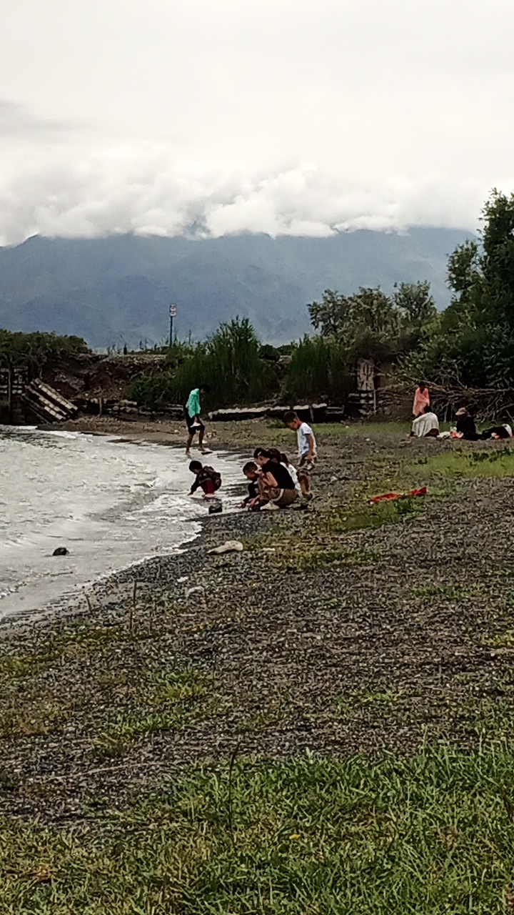 A group of people are gathered on a rocky shore by a body of water, possibly a lake, with a mountainous landscape in the background. The sky is overcast with thick clouds obscuring the peaks of the mountains. The water's edge is visible, with small waves lapping at the shore.
In the foreground, several children are playing near the water. Some are crouched down, appearing to interact with the water or the pebbles on the shore. One child in a light-colored shirt stands slightly further back, observing the others. Further up the shore, a person in a green hooded jacket stands near a wooden structure, possibly a pier or the remnants of one.
To the right side of the image, a few adults are seated on the ground, some appear to be resting or picnicking. One person is wearing a light-colored headscarf. There are some bags and blankets visible near them.
The overall scene suggests a casual outdoor gathering on a cloudy day. The presence of water, the rocky shore, and the people's attire indicate a temperate climate. The mountainous backdrop suggests a scenic location, likely in a region with natural beauty. The weather appears to be overcast, possibly hinting at a cooler or damp day. There is a signpost with a red and white circular symbol visible in the distance.