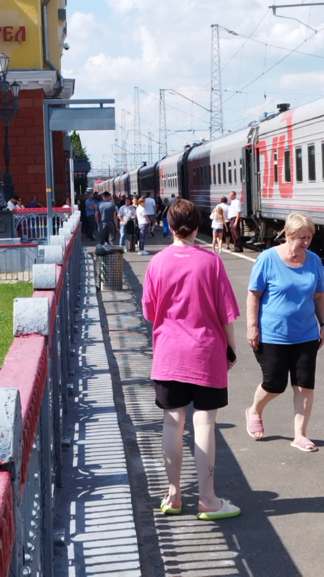 The image captures a scene at a train station in Oryol, Russia, on a sunny day with a partly cloudy sky. In the foreground, two women are walking away from the camera on a platform. The woman on the left wears a bright pink t-shirt and black shorts, with light-colored slip-on shoes. The woman on the right is dressed in a blue t-shirt and black shorts, wearing pink sandals.
Behind them, a train with silver and red carriages is stopped at the platform, with its doors open. A group of people, some waiting to board or disembark, are visible near the train. To the left of the platform, a red brick building with white trim and an awning is partially visible, featuring some yellow lettering that might be part of a sign. A lamppost is also present near this building. The platform itself is paved and features a railing along the edge. Shadows from the railing create a striped pattern on the ground. In the background, electrical wires and pylons associated with the railway are prominent against the sky.