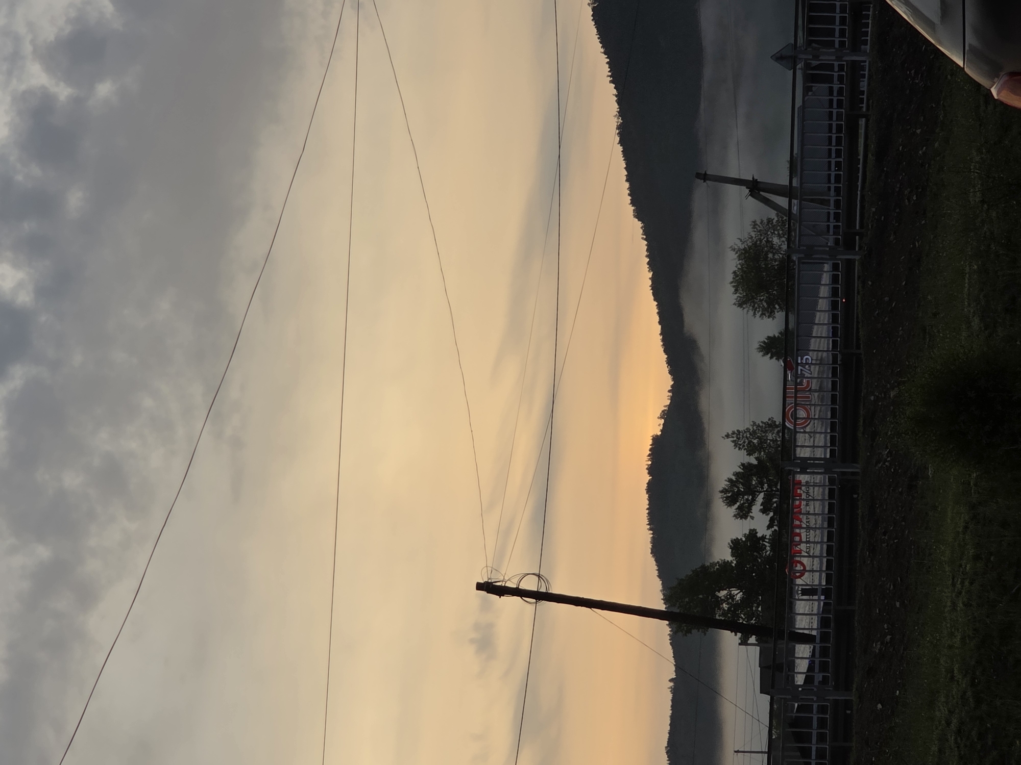 This image depicts a dimly lit outdoor scene, likely at dawn or dusk, with a soft, orange-hued sky and muted grey clouds. A power line stretches across the upper portion of the frame. In the middle ground, a dark silhouette of a forested mountain range is visible, partially obscured by a layer of fog. In front of the mountains, a metal fence runs horizontally, with several trees visible behind it. A utility pole with tangled wires stands prominently on the left side of the frame. A sign with Russian text and the number "75" is visible on a structure behind the fence. The foreground shows a dark, grassy area. The overall impression is one of a rural or semi-rural landscape with infrastructure elements.