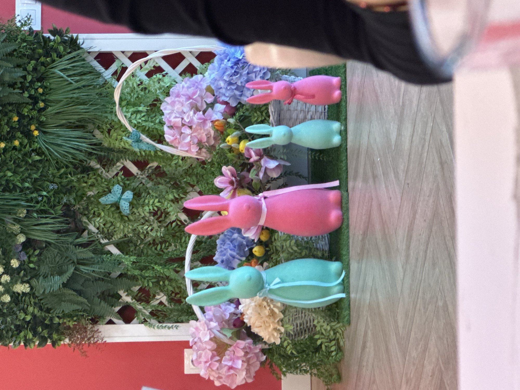 The image shows a decorative Easter display. It appears to be indoors against a red and white wall. A white lattice structure serves as a backdrop for artificial greenery. Two baskets filled with artificial flowers in shades of pink, purple, and cream are positioned against the greenery. Four pastel-colored bunny figures are placed among the flowers. Two are pink, and two are green. Each bunny has a ribbon tied around its neck. The location is Sofia, Bulgaria. A person is in the image as well but it is unclear the context of the person. The floor has a wood pattern design.