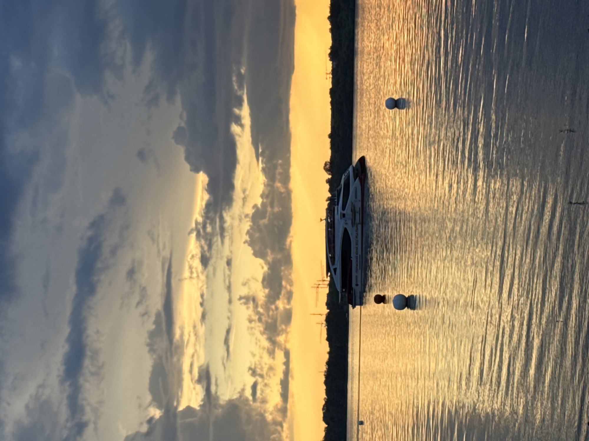 This image captures a serene sunset scene over the Kama River in Perm, Russia. A sleek, modern white and red hydrofoil boat is positioned mid-river, gently gliding on the water. The river's surface is a shimmering expanse of golden light, reflecting the dramatic colors of the sunset. Three buoys are visible in the water: one red and two white, marking a section of the river.
The sky is a spectacle of contrasting colors. Dark, heavy clouds dominate the upper portion of the frame, tinged with grey and hints of deep blue. Below these, a band of vibrant golden and yellow light stretches across the horizon, indicating the setting sun. The sunlight pierces through gaps in the clouds, creating a dramatic and atmospheric effect.
In the distance, a dark line of trees forms the bank of the river, separating the water from the sky. Above the tree line, several construction cranes are faintly visible, suggesting ongoing development in the city of Perm. The overall impression is one of tranquility and natural beauty, with the man-made elements of the boat and cranes adding a subtle touch of human presence to the landscape. The water's surface is rippled, creating a dynamic texture that complements the stillness of the boat and the vastness of the sky.