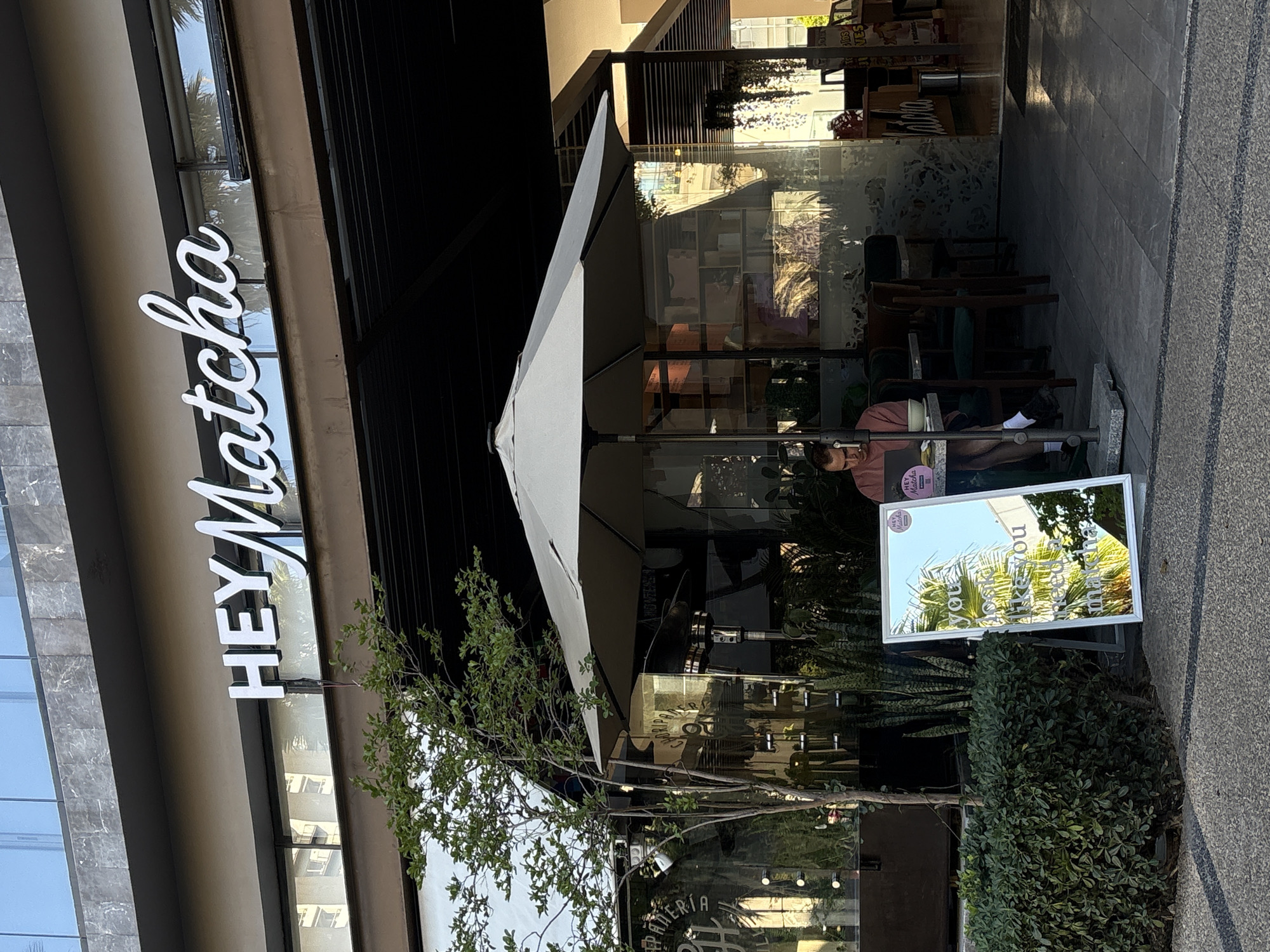 A daytime outdoor scene shows the entrance of the "HEY Matcha" cafe in San Bernardino Tlaxcalancingo, Mexico. The white "HEY Matcha" sign is prominently displayed vertically on the building's facade. An awning covers outdoor seating, where a person sits at a table. A sign displaying "you can take matcha" is visible at street level among foliage. The facade of the modern building includes mirrored glass sections. The setting suggests a business area on a sunny day.