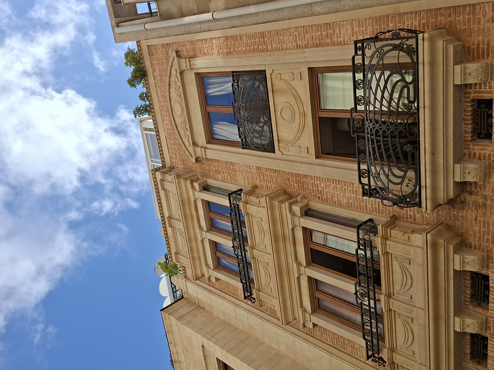 The image shows a close-up, low-angle shot of a building facade in Torrevieja, Spain. The building has multiple stories with windows and balconies. The facade is constructed with a combination of light brown stone and reddish-brown brick. The windows have wooden frames and some have white curtains visible. There are decorative wrought-iron balconies with intricate patterns on the upper floors. The architectural style appears to be a blend of classical and modern elements. The sky above is blue with scattered white clouds. A portion of an adjacent building, made of grey concrete, is visible on the right. Small green plants are seen on the rooftop. The scene depicts a residential or commercial building in an urban environment.