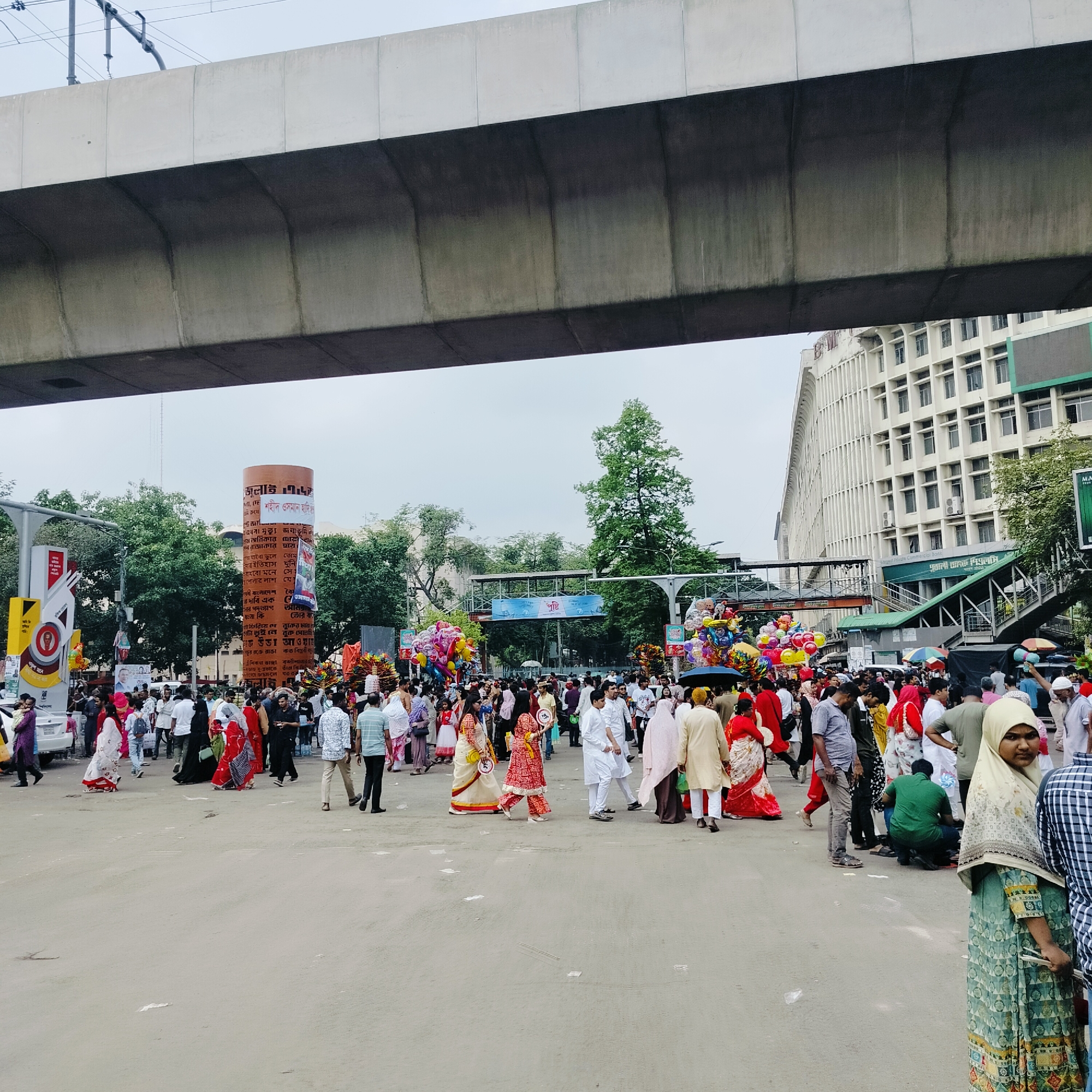 The image captures a crowded street scene in Dhaka, Bangladesh, possibly during a festive or commemorative event. A large group of people, many dressed in traditional attire such as saris and punjabis, are gathered in a large open area beneath an elevated concrete structure, possibly a flyover or metro line. A tall, cylindrical monument stands to the left of the frame, adorned with Bengali script and posters. The architecture in the background includes multi-story buildings. Trees and a pedestrian overpass are visible in the midground. Several individuals appear to be carrying balloons, and street vendors may be present amid the crowd. The overall atmosphere suggests a public gathering or celebration taking place in a busy urban setting.