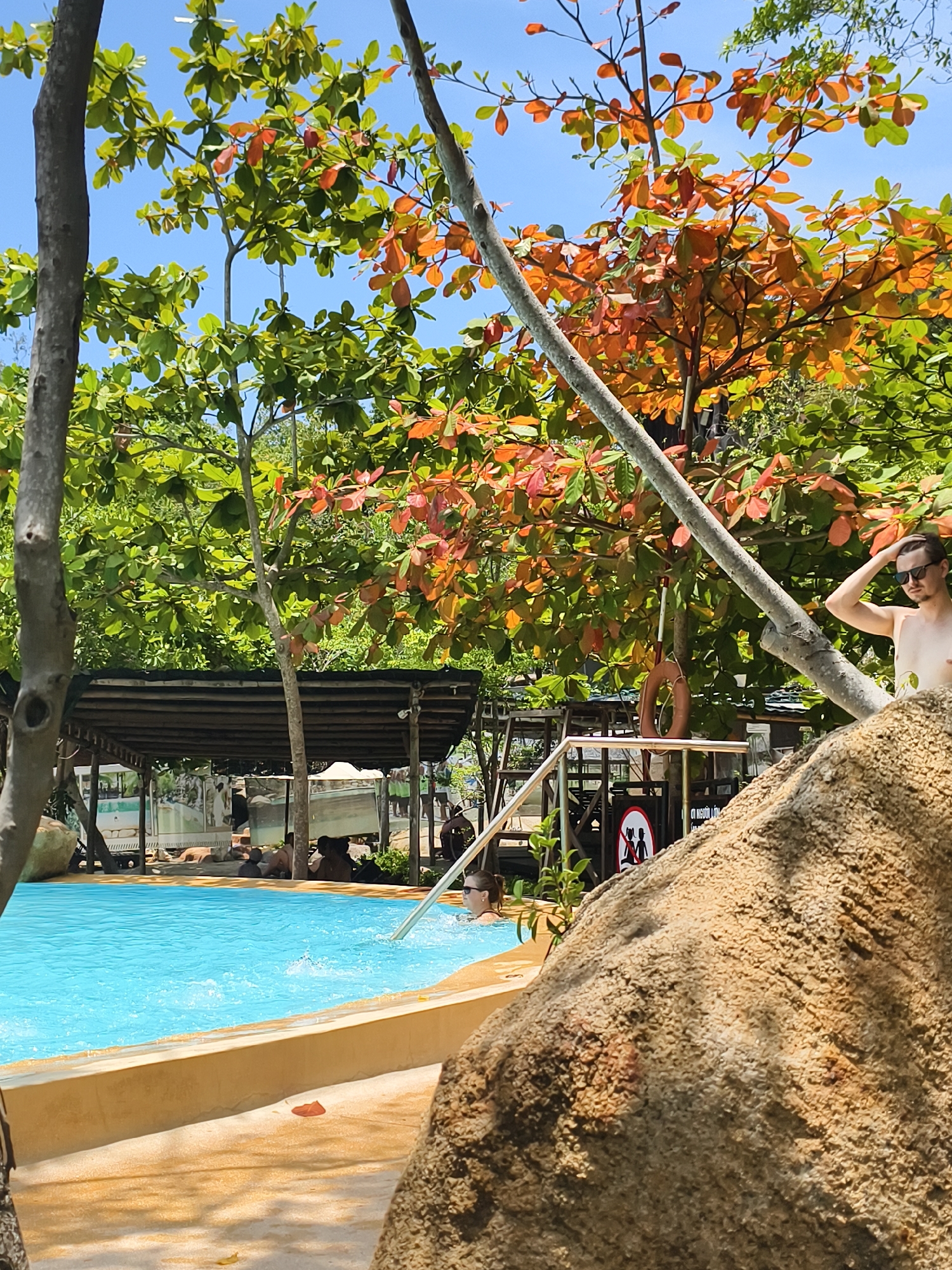Captured in Tay Nha Trang Ward, Vietnam, the image presents a poolside scene on a sunny day. A turquoise pool is bordered by a light-tan concrete edge. In the pool, a woman swims, her head visible above the water. Around the pool's perimeter, several people are seated or lying under a covered structure with a dark roof. A partially obstructed man with sunglasses stands near a large, textured rock formation. The vegetation consists of green and red-orange trees that fill much of the background. A tree branch cuts diagonally across the frame. A safety ring hangs on a metal railing next to a sign featuring stick figures.