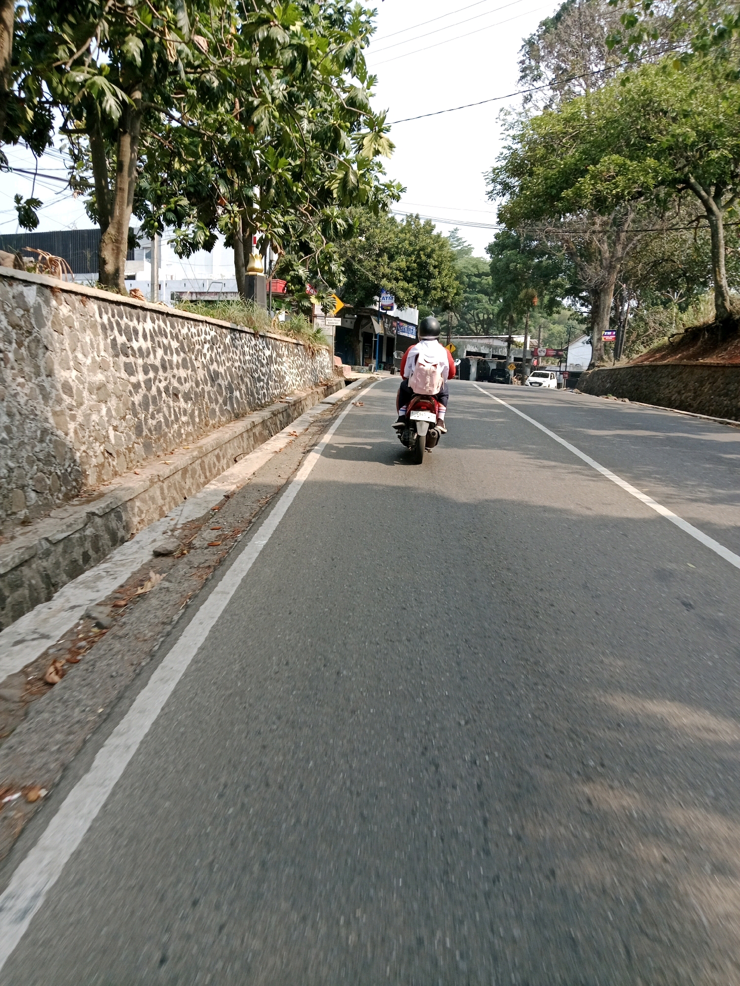 The image shows a person riding a scooter on a paved road in Sirnagalih, Indonesia. The rider, wearing a white helmet and a red and white jacket, has a light pink backpack on their back. They are traveling on the right side of the road, which has a white line marking the lane. The road is bordered on the left by a stone wall with a drainage ditch. Lush green trees line the right side of the road, and some buildings are visible in the background. The sky is bright, indicating daylight.