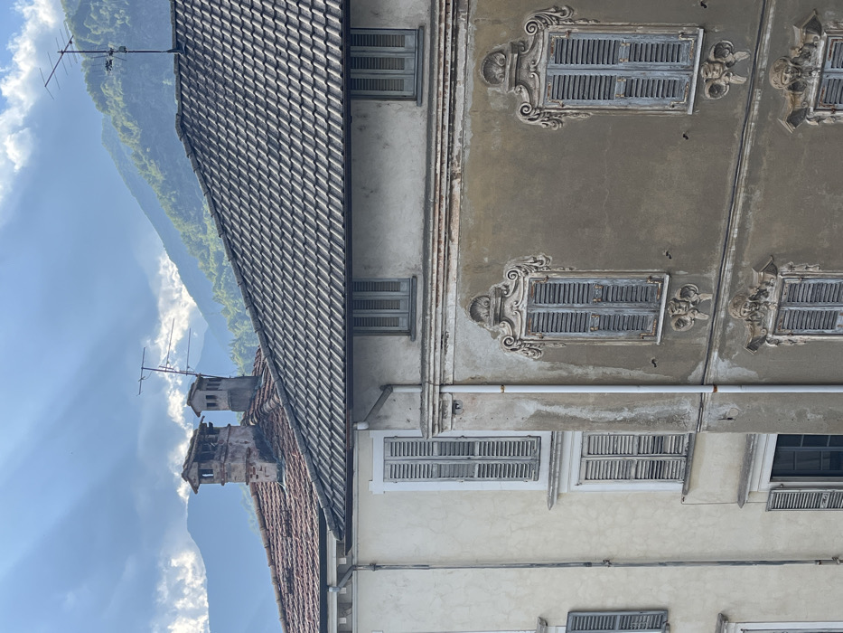 This image depicts a detailed view of architectural facades and rooftops in Varallo, Italy, set against a mountainous backdrop under a partially cloudy sky. In the foreground, two buildings are prominently featured. The building on the right has a dark grey tiled roof, with two small rectangular windows on its upper floor, fitted with light blue-grey closed shutters. Below this, its main facade is a weathered brownish-grey, showcasing two rows of windows. Each window is framed by elaborate, ornate, weathered light grey stucco decorations in a Baroque style, incorporating shell motifs at the top and sculpted faces or cherubs at the bottom. All windows on this section are fitted with light blue-grey closed shutters. A white vertical downspout runs along the left edge of this building's facade.
To the left, a portion of a lighter cream-colored building is visible, topped with a red-orange tiled roof from which two weathered brick chimneys protrude. Multiple windows with simple light blue-grey closed shutters are visible on its facade.
Above the rooftops, two television antennas are mounted. In the background, hazy, green, tree-covered mountains rise. The sky is predominantly blue with scattered white and grey clouds, from which distinct sunbeams, or crepuscular rays, are visible radiating downwards. The scene is illuminated by natural light, casting subtle shadows.