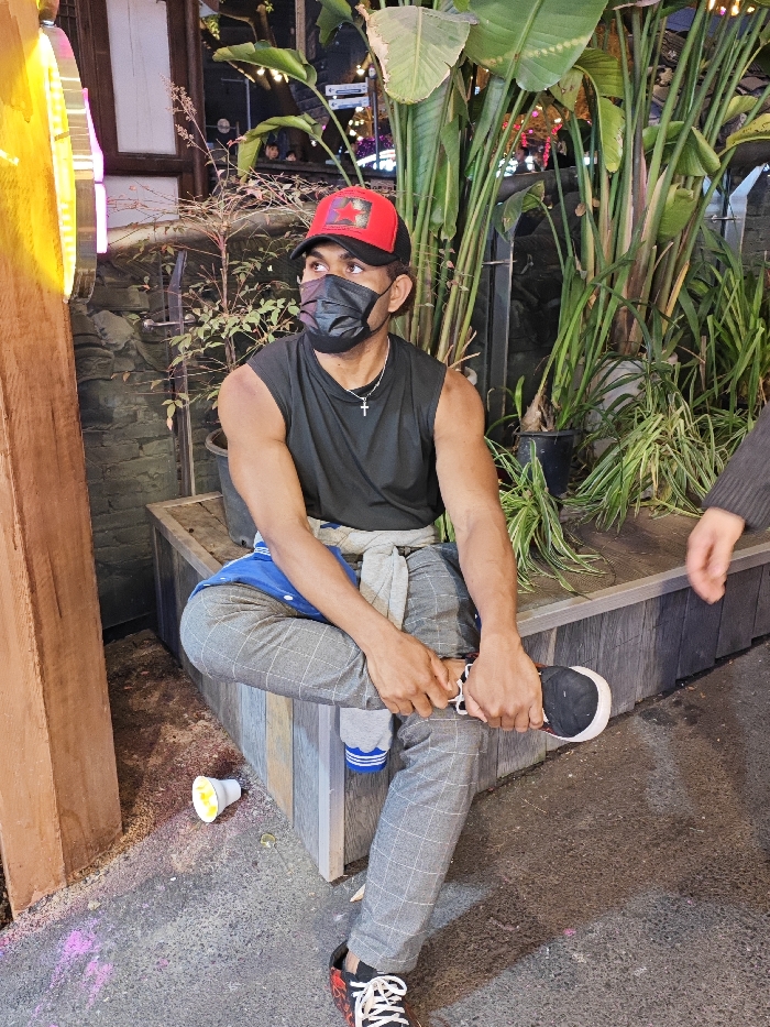 This image captures a male individual seated outdoors at night in Yuzhong District, China. The subject, of darker complexion, is depicted wearing a black face mask, a black sleeveless shirt, grey plaid trousers, and a red and black baseball cap featuring a red star emblem. A silver cross pendant hangs from his neck. A grey sweatshirt is tied around his waist, with a blue garment visible beneath it. He is positioned with his right leg crossed over his left, his hands clasped around his left foot, which is shod in a black and red sneaker. White socks with blue stripes are visible on his left ankle. He is seated on a multi-toned wooden slatted bench. To the left, a tall wooden beam and a vertical yellow neon light fixture are present. The background features several large potted plants with broad green leaves, a stone wall, and illuminated string lights, suggesting a commercial or recreational area. The ground surface is dark and paved, with scattered pink and yellow powdery residue and a discarded white paper cup with a yellow interior lying near the subject's left foot. A lighter-skinned hand, clad in a dark sleeve, is partially visible on the far right edge of the frame, near the bench.