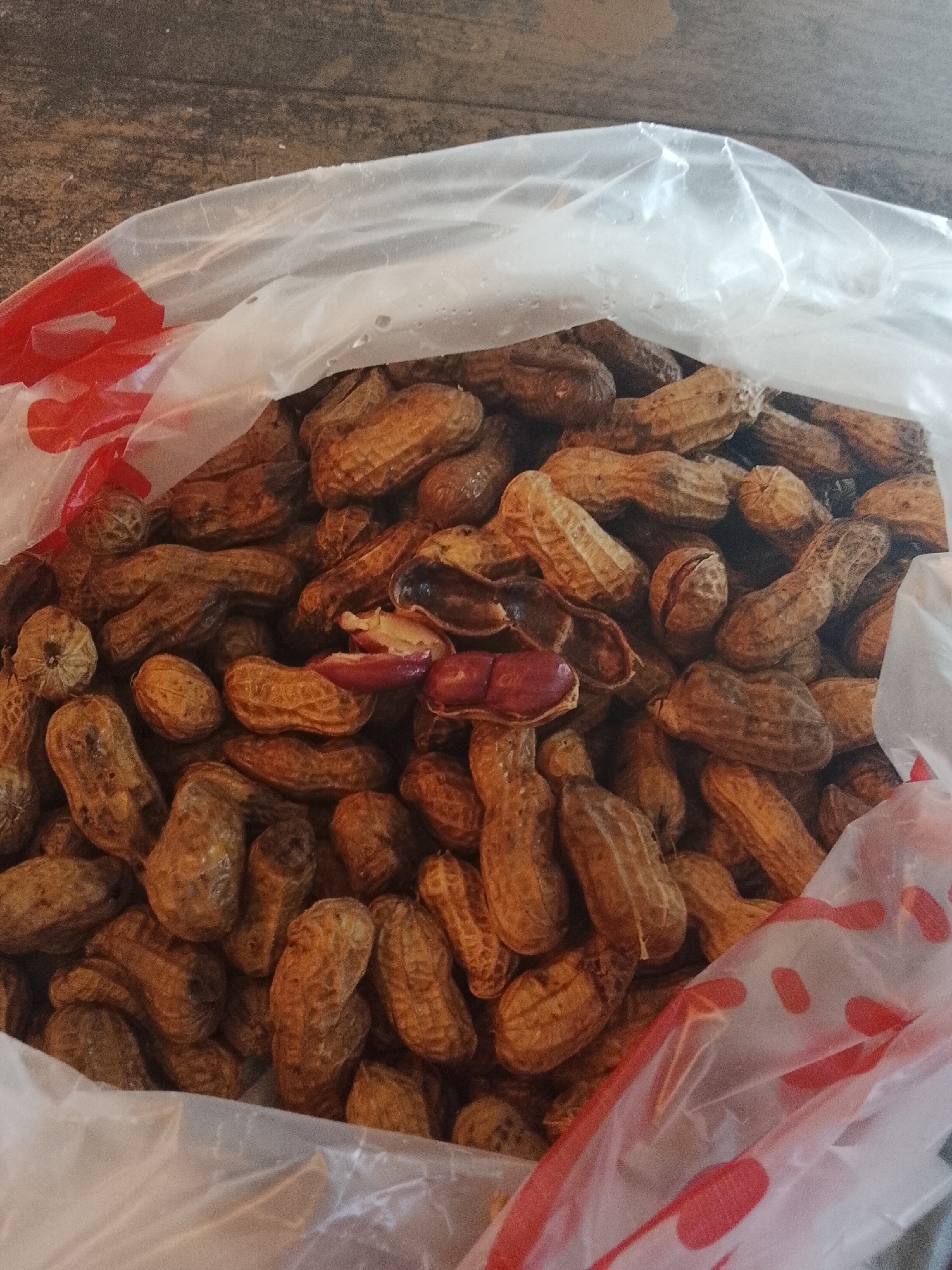 A pile of unshelled peanuts, some cracked open to reveal the red-skinned nuts inside, are contained within a clear plastic bag with a red polka-dot pattern near the top edge. The bag is resting on a wooden surface. The image is a close-up shot, focusing on the texture and arrangement of the peanuts. No people or specific city features are visible.