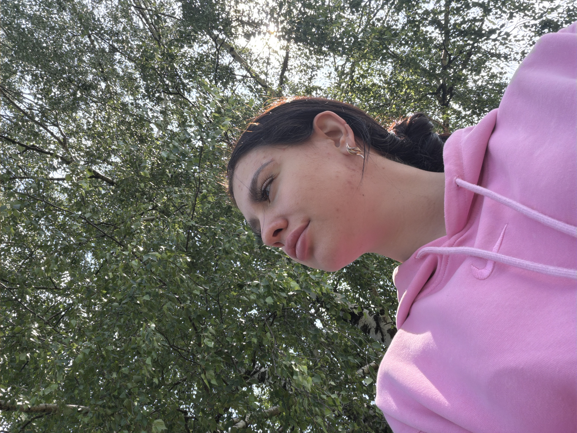 A medium shot shows a young woman wearing a pink hoodie, looking up through a canopy of green leaves. The camera angle is from below, looking upwards towards the sky. The woman's face is visible in profile, with her eyes looking upwards. Her hair is dark and pulled back. She has a small earring in her left ear. The leaves of the trees are dense and partially obscure the bright sky, with sunlight filtering through the branches. The background consists of trees and sky, suggesting an outdoor setting. The image does not contain any specific details indicating a location in the city of Чумна, Russia.