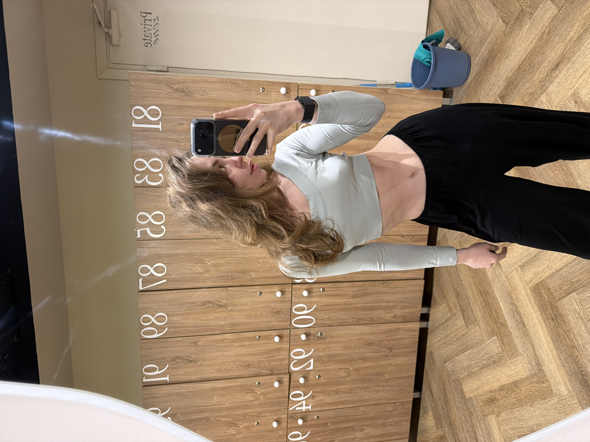 A woman is taking a selfie in a locker room. She is wearing a light green long-sleeved crop top and black sweatpants. She is holding a smartphone in her left hand. The locker room has wooden lockers with white numbers on them. A blue bucket with cleaning supplies is visible in the background. The location is Moscow, Russia.