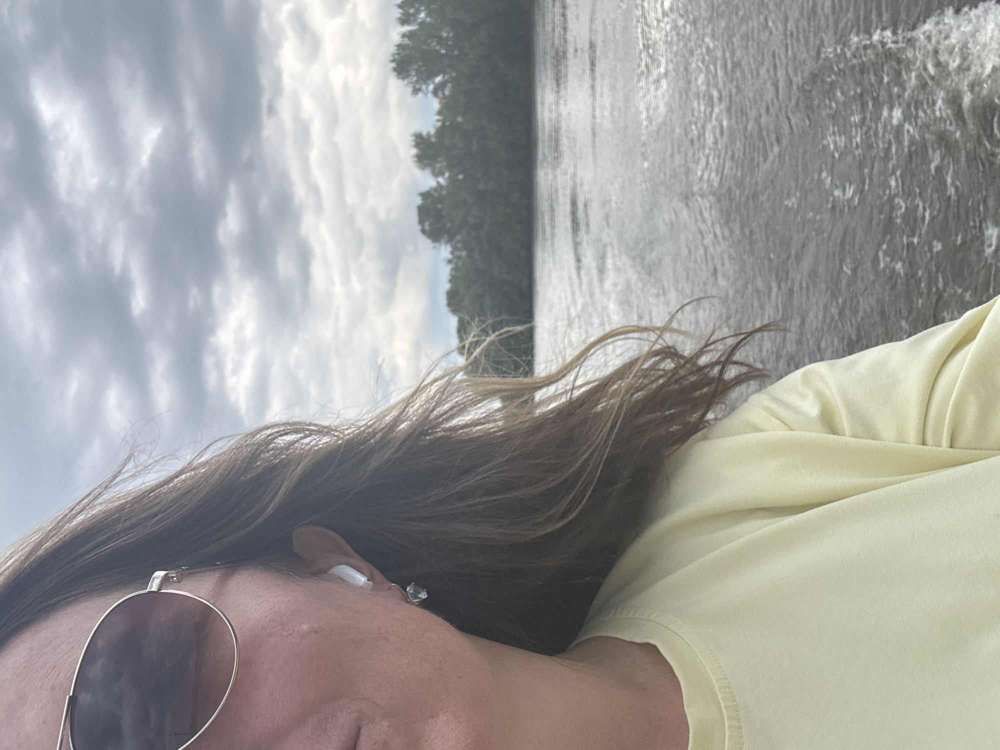 A close-up, half-profile shot of a woman with long, brown hair and sunglasses is taken from a boat on a river. She is wearing a light yellow sweatshirt and a white earbud in her left ear, along with a dangling earring. The river is choppy with a wake behind the boat, and a tree line is visible on the far bank under a cloudy sky. The image does not contain enough information to determine the location within the city of Ufa, Russia.