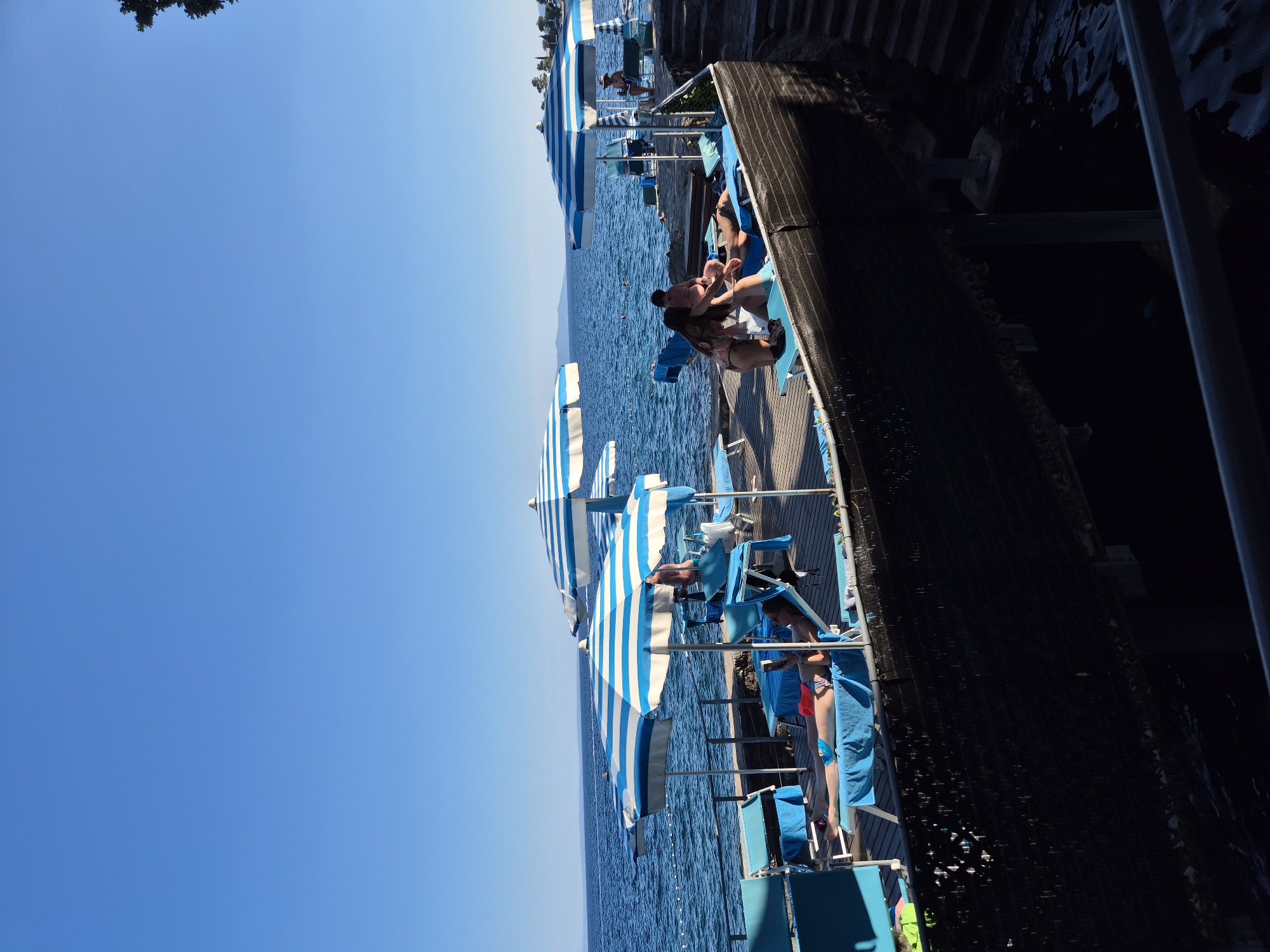 In an outdoor scene on a sunny day, a group of people relax on a wooden deck near the water. The deck is lined with blue and white striped umbrellas providing shade over lounge chairs. Some individuals are seated or lying on the chairs, with one person reading. Other people are standing or interacting with each other near the lounge chairs. The water, a deep blue, is visible beyond the deck. A few trees are visible in the distance on the far shore. The scene appears to be a resort or public beach area.