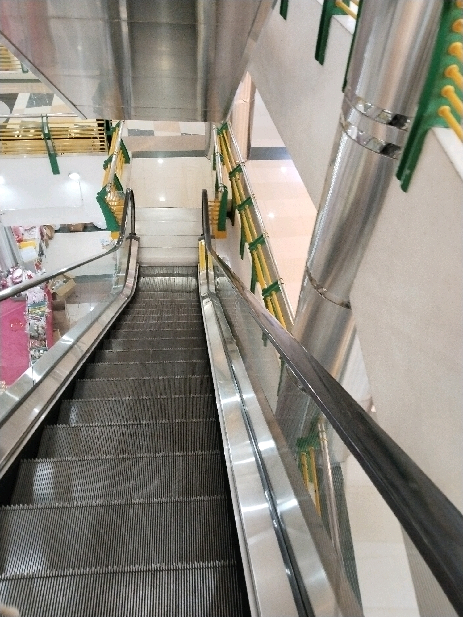 An overhead view shows a downward-sloping escalator in a mall located in Surabaya, Indonesia. The escalator's metal steps are visible, transitioning into a polished metal tread at the bottom. To the right, a section of the mall's interior is visible, featuring white walls and a portion of a store with shelves displaying various items. The escalator is enclosed by glass panels on both sides. A stainless steel handrail runs along the top of the glass, and the escalator is framed by green and yellow railing supports. The floor tiles on the right appear to be a light beige.