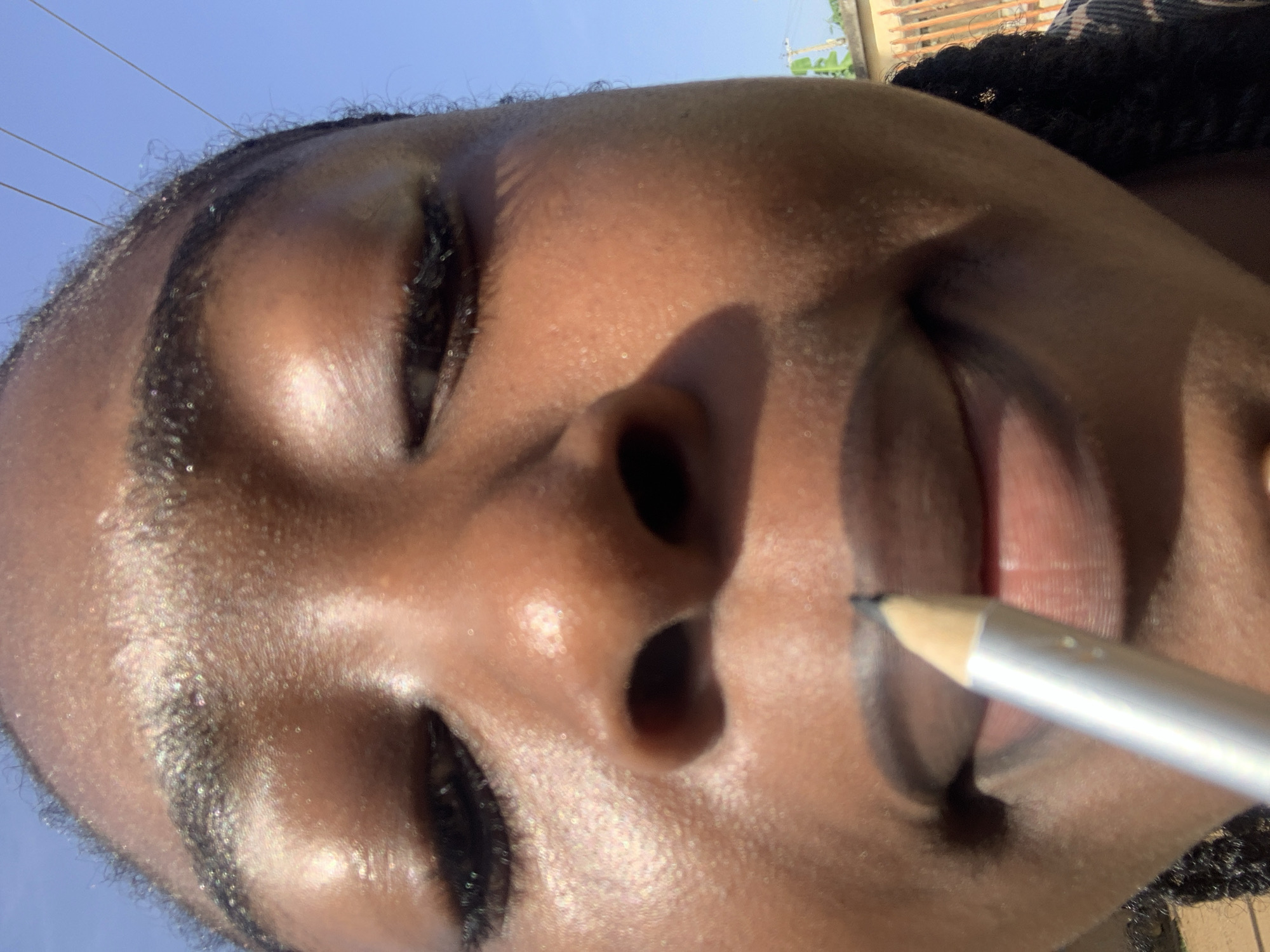 An upward-facing close-up shot captures a person with dark skin applying makeup. Their eyes are closed in concentration as a light-colored pencil, possibly an eyeliner or lip liner, is held to their upper lip. Strong, direct sunlight illuminates the person's face, creating distinct shadows and highlighting the natural sheen on their skin, which shows visible texture and pores. Braided hair is visible along the edge of the frame, and their eyebrows are well-defined. The background is dominated by a clear, bright blue sky with faint horizontal lines, likely power lines, indicating an outdoor setting. The intense illumination suggests it is sunny daytime, possibly midday or early afternoon. This scene is set in Rafin Par, Nigeria. There is no visible text in the image.