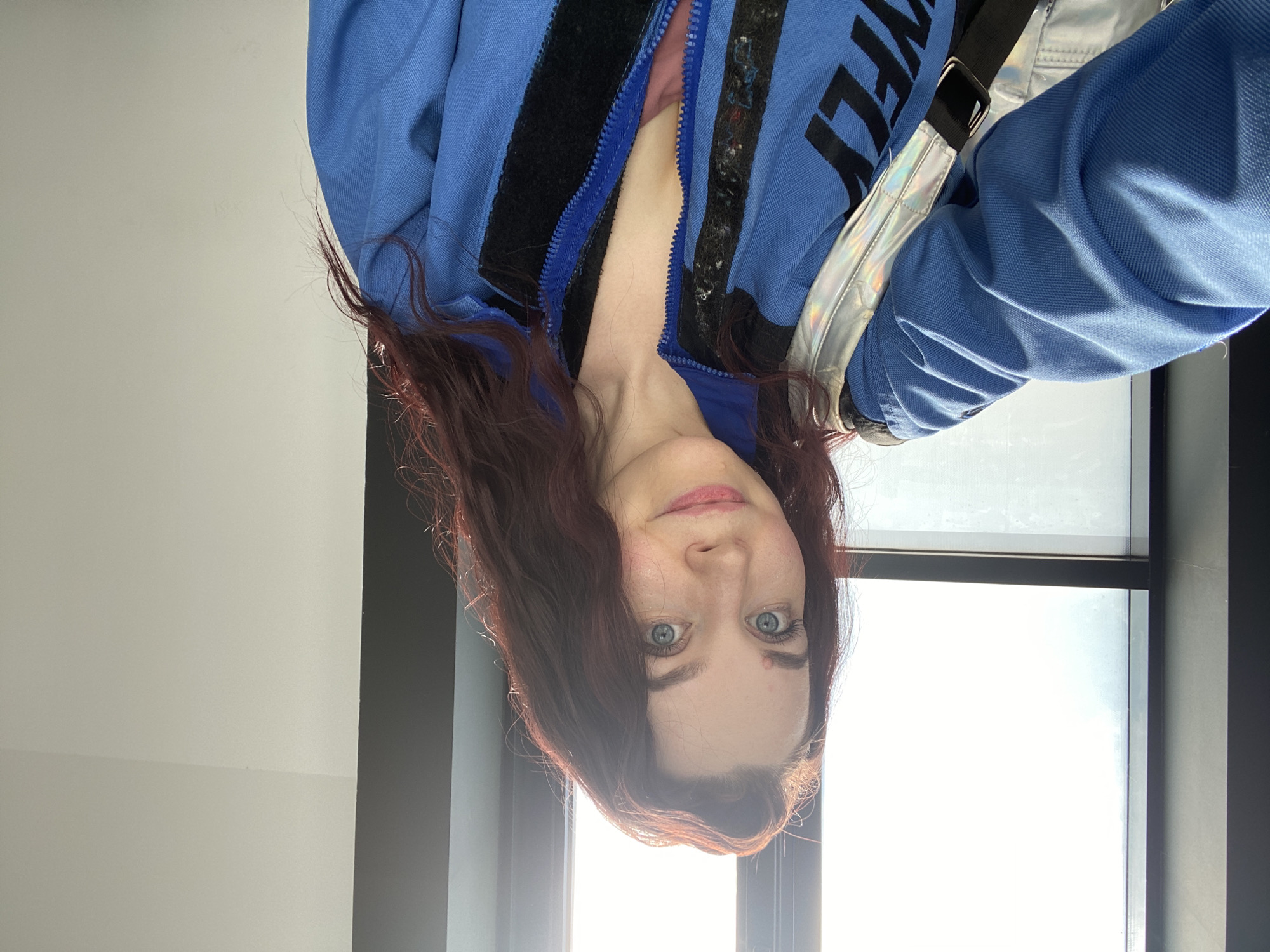 A fair-skinned woman with red-brown hair stands in front of a bright window. The photo is oriented so that the woman's head is at the bottom of the frame. She is wearing a blue jacket with a zipper partly open and black stripes. The jacket has the word "FLY" visible on the right side. A silver strap is visible over her shoulder. The wall to the left is white, while the frame around the window is dark gray. It is not possible to verify the location in the city of Izhevsk, Russia, from the image alone.