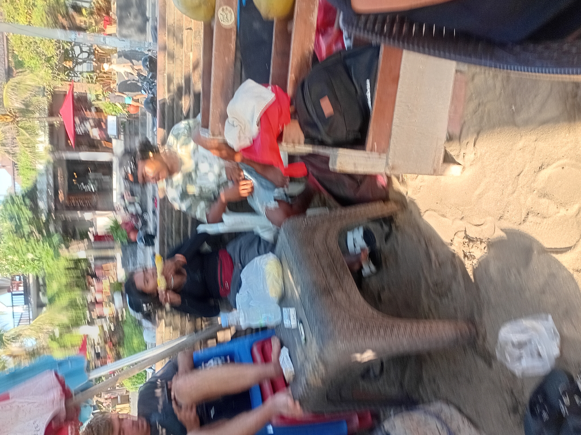 In Kuta, Indonesia, a group of people are gathered on a sandy beach area with tiered seating constructed of wood and stone steps in the background. A woman in the foreground is eating corn on the cob, while a man sits to her left, looking down. To the right, another man wears a camouflage shirt and appears to be resting. Various items are scattered around, including a plastic table with a water bottle and snacks, bags, and clothing. Beyond the seating area, a building with a red umbrella and greenery suggests a restaurant or cafe. The scene appears to be a casual outdoor gathering, possibly at a beachside establishment.