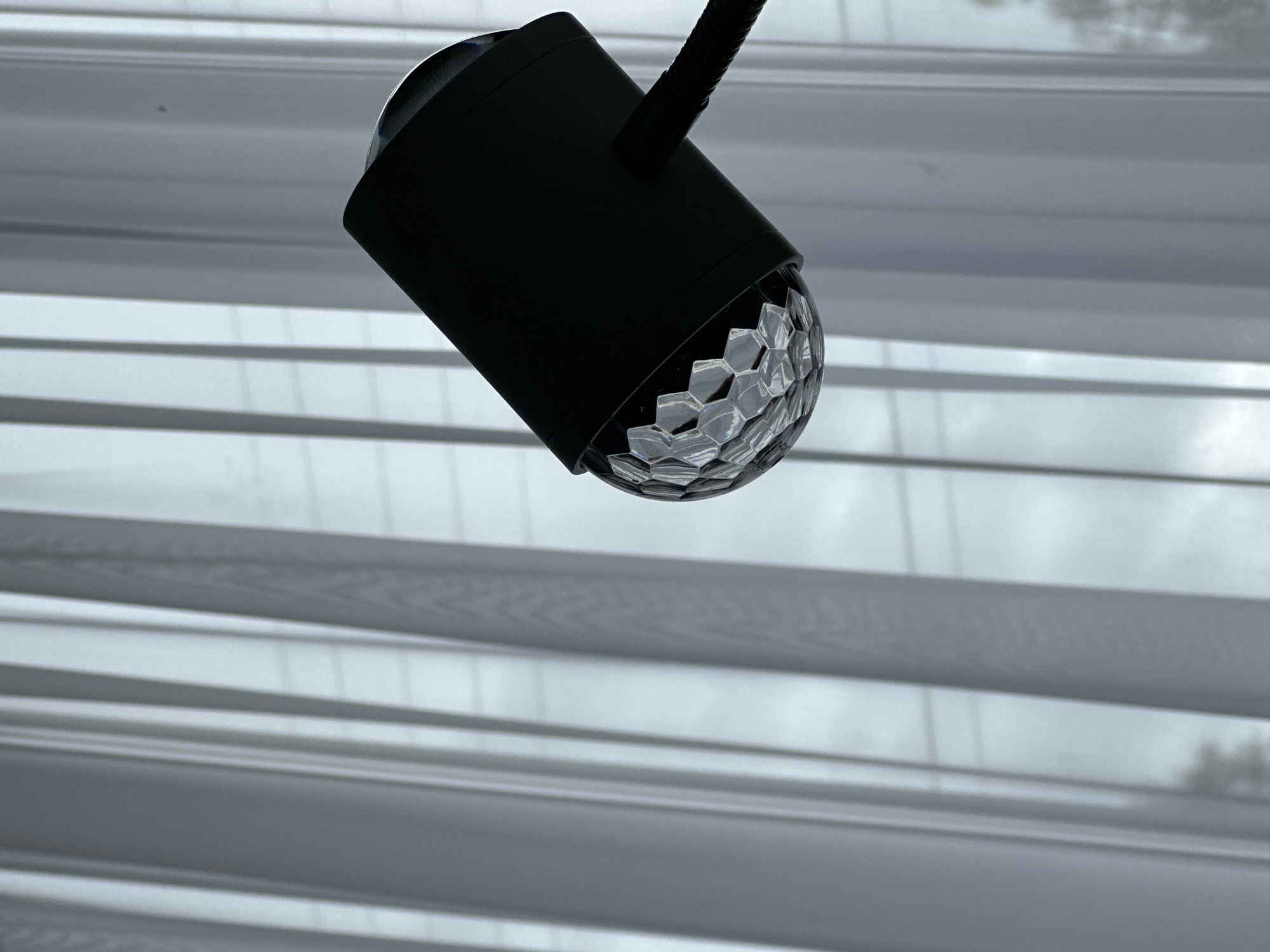 A black, cylindrical lamp with a faceted, semi-transparent dome at its lower end is mounted on a flexible arm. The lamp is positioned against a background of horizontal blinds, which are open and allow a view of a blurred exterior scene. The location is Unknown, Unknown.
