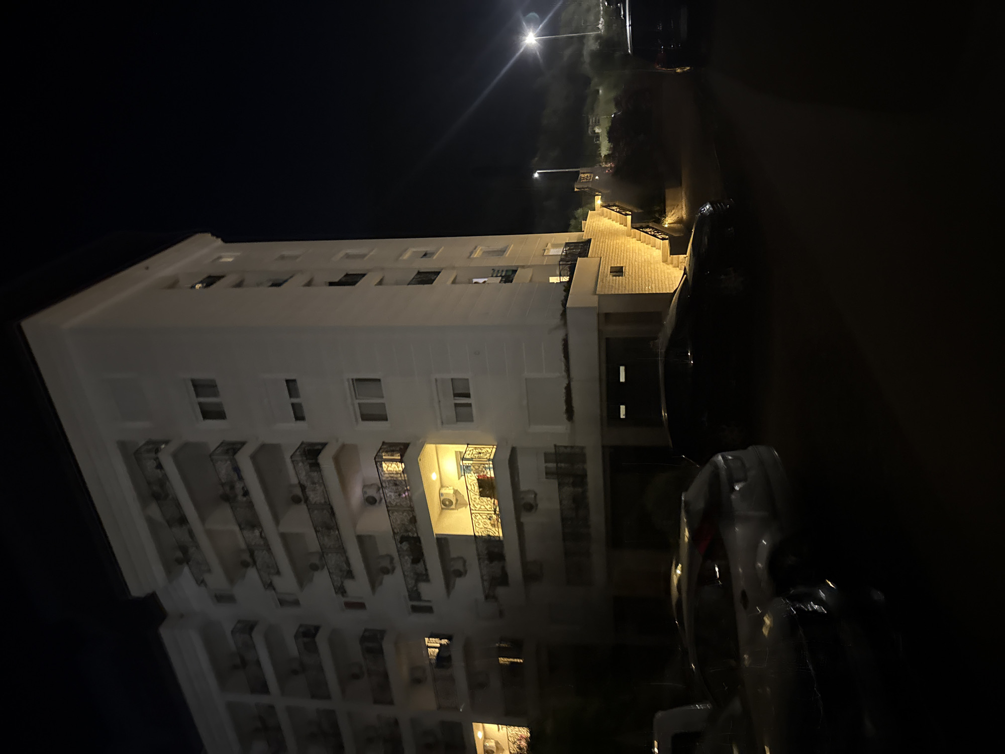 The image depicts an apartment building at night, located in Boreti, Montenegro. The building is white and has multiple balconies with dark railings. Some of the balconies have lights on, illuminating the facade. Air conditioning units are visible on the exterior walls. A set of illuminated stairs leads up to the building's entrance. Several cars are parked in front of the building on a paved surface. The background is dark, with a bright streetlamp visible in the distance and a hint of trees or foliage on a slope. The overall scene suggests a residential area at night.