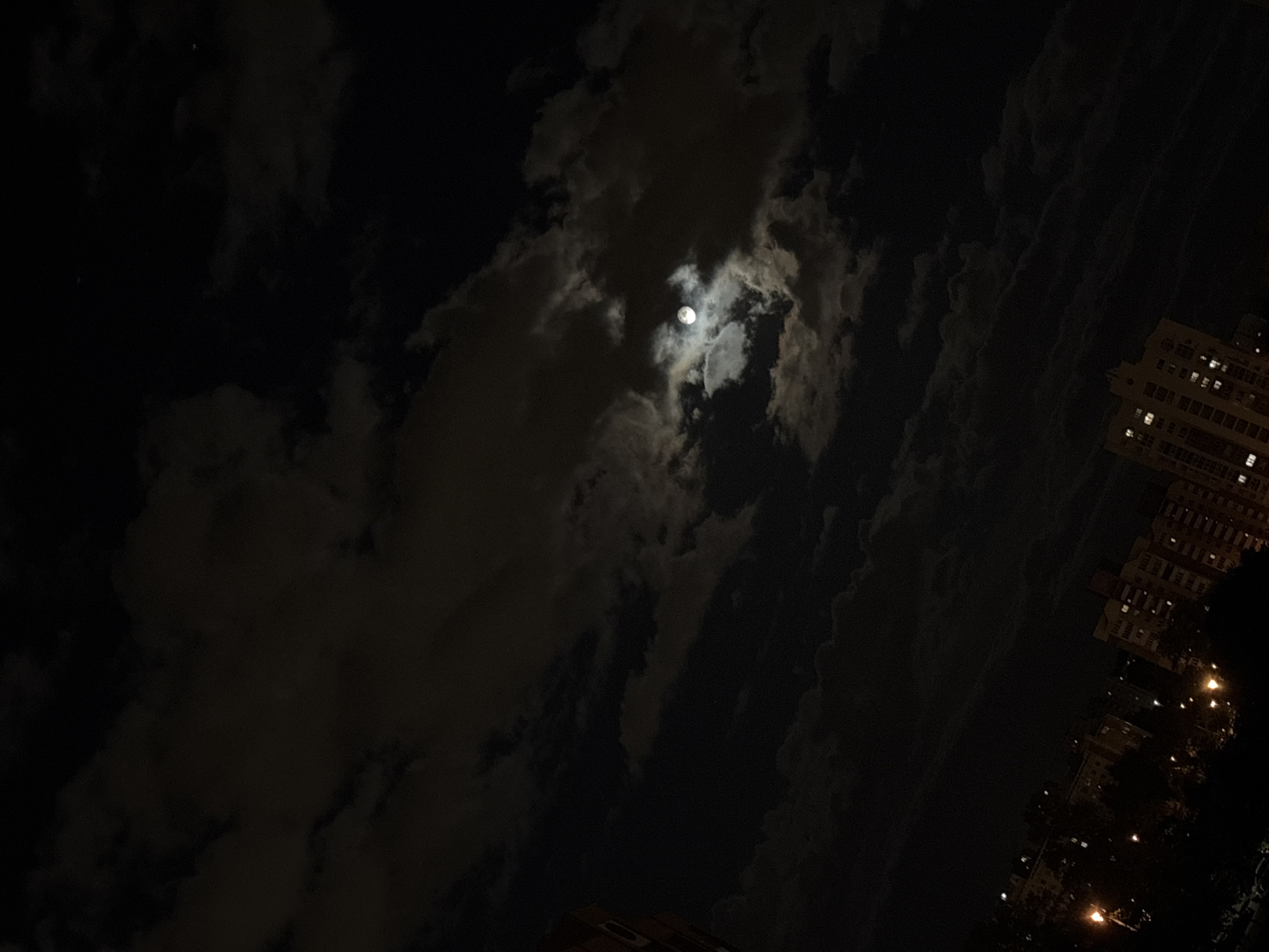 The image captures a night scene, likely in an urban environment. The primary subject is the moon, partially obscured by clouds, illuminating the surrounding sky. To the right of the frame, the lit windows of a building are visible, indicating an inhabited structure. Below the building, faint lights suggest streetlights or ground-level illumination. The overall impression is a low-light scene with a focus on the interplay between the moon, clouds, and urban architecture. There is no evidence to suggest that the location is Minsk, Belarus.