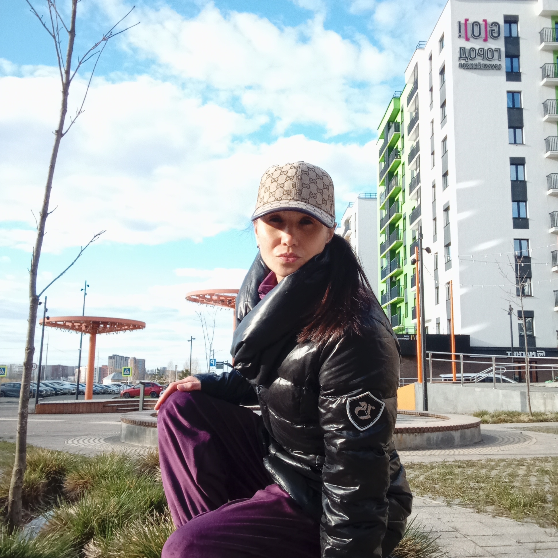 A woman is crouching outdoors in Pervomaysky, Russia, on a gray paved area adjacent to a low, circular bricked structure with tufts of grass growing around its base. The woman is wearing a brown cap with a designer pattern, a shiny black puffer jacket, and purple velvet pants. She is looking directly at the camera.
Behind her is a modern apartment complex with white walls and alternating green and gray balconies. The complex has signage in Russian on its upper floors. To the left of the woman are orange, disc-shaped structures on tall posts and, in the background, a distant cityscape with cars and buildings. The sky is partly cloudy. A thin tree trunk stands to her left.