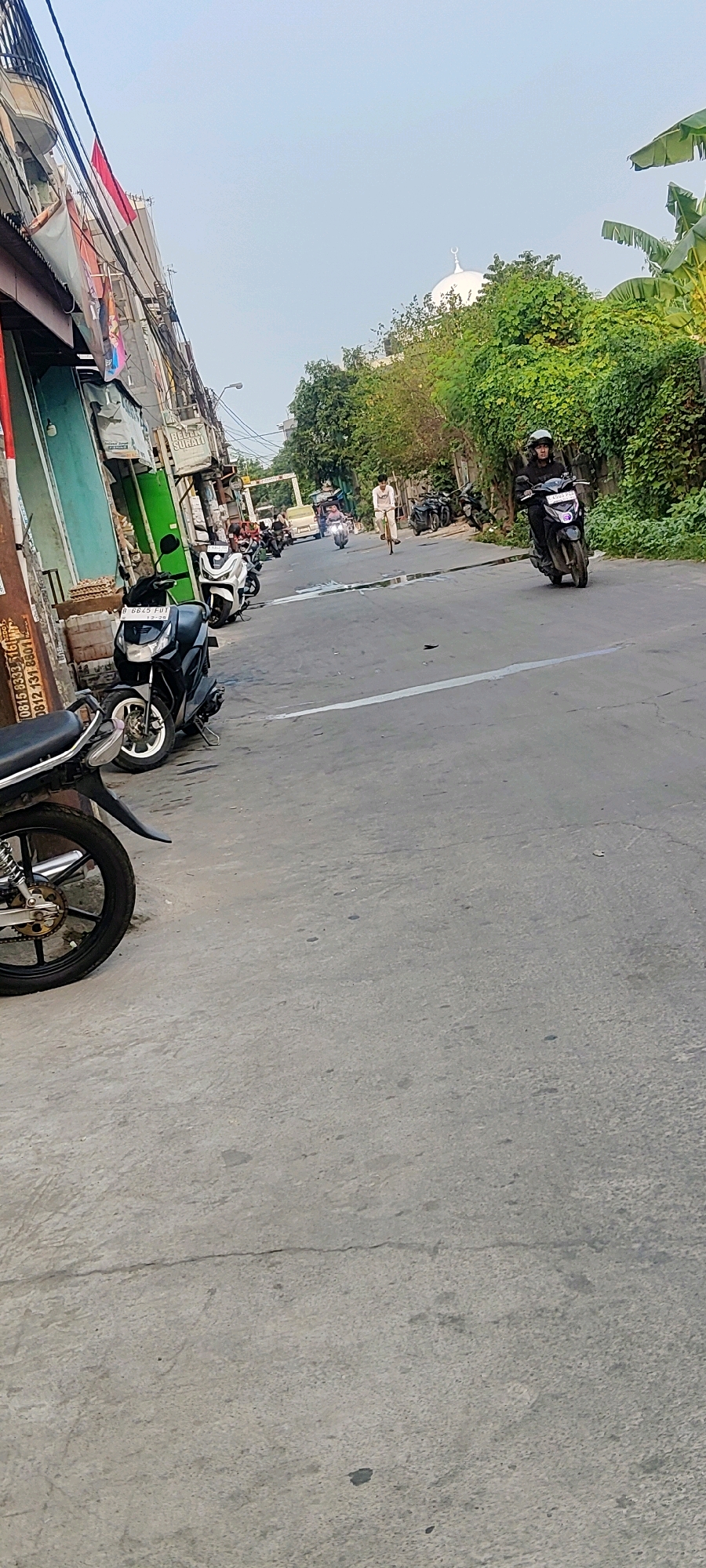 The image captures a street scene in Mekarmukti, Indonesia, with a focus on motorbikes and a mosque in the background. In the foreground, a black motorbike is parked on the left side of the road. The road itself is paved with concrete and shows some signs of wear, including cracks and water stains. Further down the road, several other motorbikes are parked and in motion. A person on a bicycle is also visible, riding down the street.
In the distance, a white mosque with a dome and a crescent moon symbol can be seen above a lush green canopy of trees. The sky is clear and bright. The overall environment suggests a typical urban or suburban setting in Indonesia. The shops on the left side of the street, with their signage and awnings, indicate a commercial area. The presence of parked and moving vehicles, including motorbikes and a bicycle, highlights the common modes of transportation in the area.