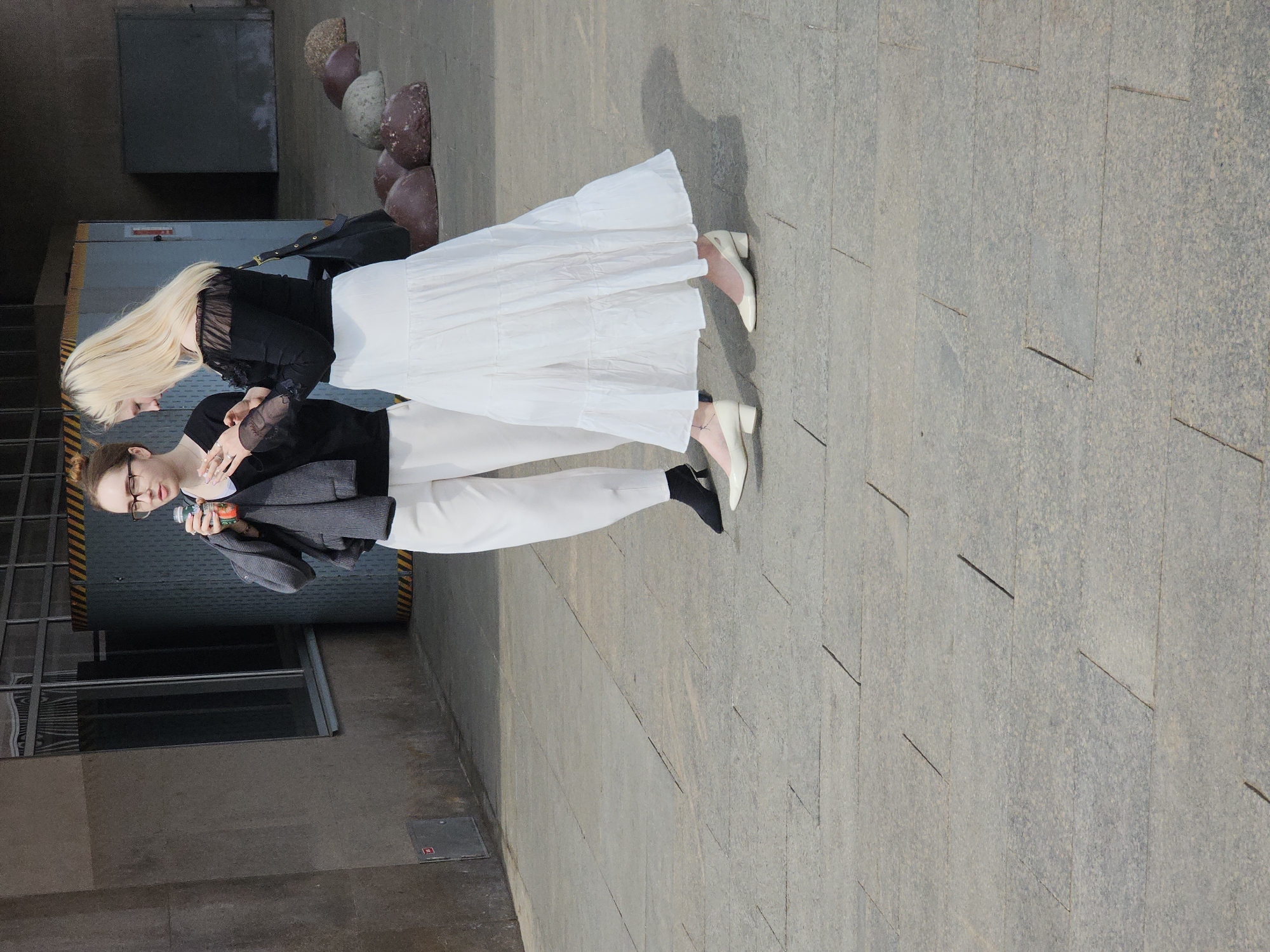 Two women stand on a paved surface, likely in an urban area in Moscow, Russia. One woman has blonde hair and wears a black top and a white skirt. The other woman has glasses, brown hair styled in a bun, wears a black top, white pants, and a grey jacket. She holds a drink. The location appears to be near a building with grey walls and a door or gate with yellow and black striped accents. There are decorative stone spheres behind them. The paved ground is grey and consists of rectangular slabs.
