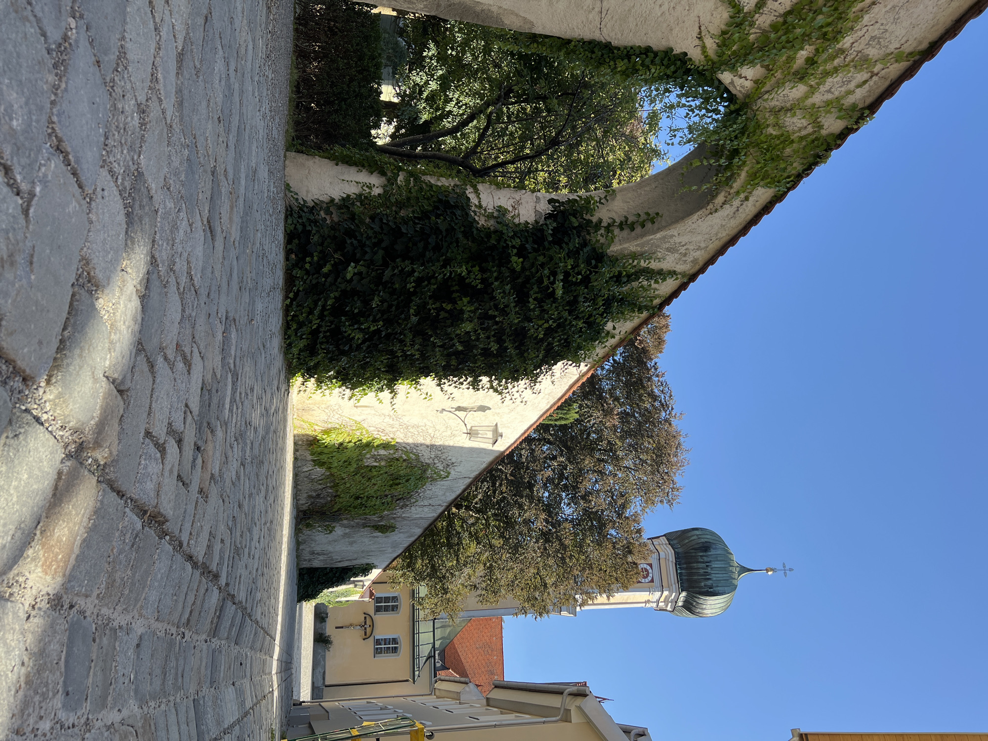 The image depicts a cobblestone alleyway in Immenstadt im Allgäu, Germany, on a clear, sunny day. A large stone archway covered in ivy dominates the left side of the foreground, leading the eye towards a narrow street. Beyond the arch, a high, white wall extends into the background, also partially draped with ivy. A lamppost is mounted on this wall.
In the distance, a church tower with a green, onion-shaped dome and a cross at its apex rises above a cluster of trees. The church building itself, painted in a pale yellow, features a red tile roof and a crucifixion scene mounted on its facade. Other buildings with red roofs line the street to the right. The cobblestones in the alleyway are uneven, with some areas showing signs of wear. The overall impression is one of historic European architecture bathed in bright sunlight, with a strong emphasis on the textures of stone, ivy, and cobblestones. There are no people visible in the scene.