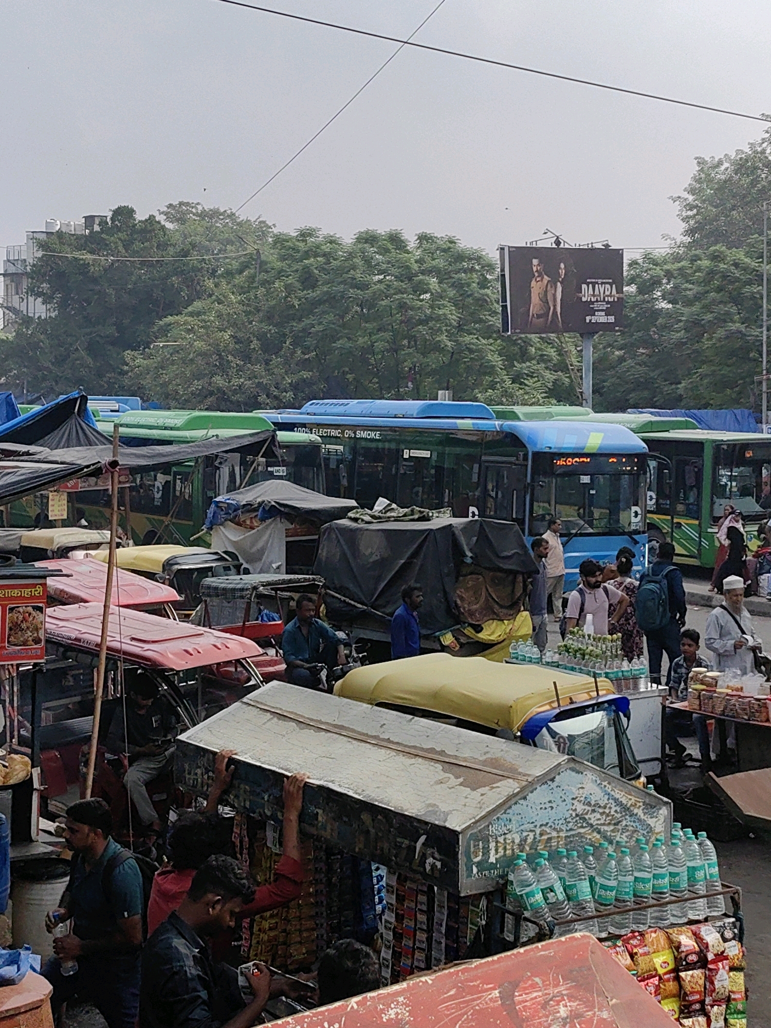 The image depicts a busy street scene in Delhi, India, likely a bus station or a busy intersection. A large number of vehicles are present, including several buses of varying colors (green and blue), auto-rickshaws, and what appears to be a yellow-hooded vehicle, possibly another auto-rickshaw or a similar mode of transport.
People are visible throughout the scene, engaged in various activities. Some are standing and waiting, while others appear to be passengers or drivers. Several individuals are interacting with each other, suggesting a bustling public space. There are also street vendors present, with carts or stalls displaying goods such as bottled water, snacks, and other items. The environment appears somewhat cluttered, with tarps and coverings over some vehicles and stalls. Trees are visible in the background, and the sky is overcast. A billboard advertising a film titled "DAAYRA" is also prominent in the upper portion of the image. The overall impression is one of a lively, crowded, and dynamic urban environment.