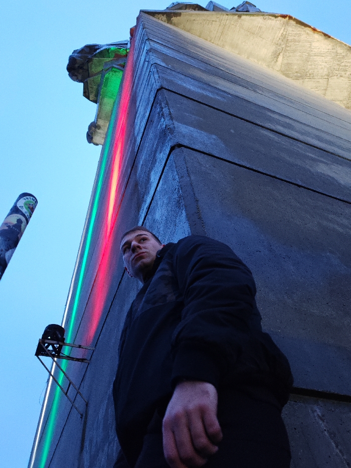 An outdoor, low-angle photograph captures a male individual standing in front of a prominent concrete structure, identified as the Monument to 1300 Years of Bulgaria, in Shumen, Bulgaria. The subject, a male in his 20s or 30s with short hair, is positioned in the lower-center of the frame, wearing a dark, padded jacket and dark trousers. He is looking upwards and slightly to his left. His right hand is partially visible in the lower-right quadrant, appearing loosely clenched. The monument, constructed from unpainted grey concrete with visible seams and sharp angles, dominates the background. Vertical strips of red and green light illuminate a section of the monument's facade. A dark-colored spotlight fixture is mounted on a metal bracket attached to the concrete wall, positioned to the left of the subject. A tall, slender pole, covered with various stickers or graffiti, is partially visible on the far left. The sky above is a clear, light blue, suggesting late dusk or early morning conditions.