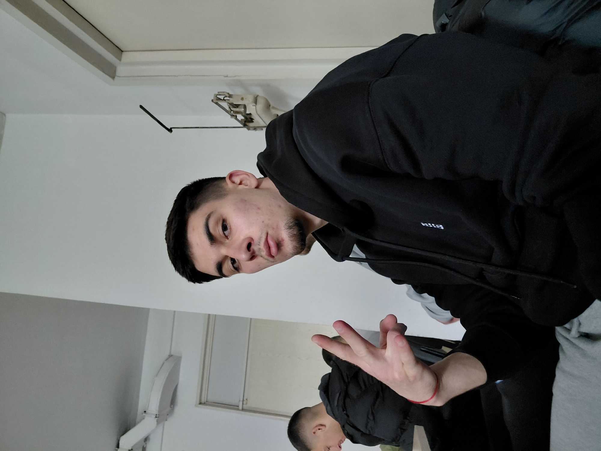 A young man in a black hoodie is posing indoors. He has short dark hair, a beard, and some acne. He is holding up three fingers in a peace sign gesture. A mirror is reflecting a similar scene, showing another person in a black jacket, likely in the same room. The room has white walls, a white door, and a vintage-style metal hanger on the wall. A hand sanitizer dispenser is visible on the mirrored wall. The location is likely indoors. Sofia, Bulgaria, is not clearly indicated.