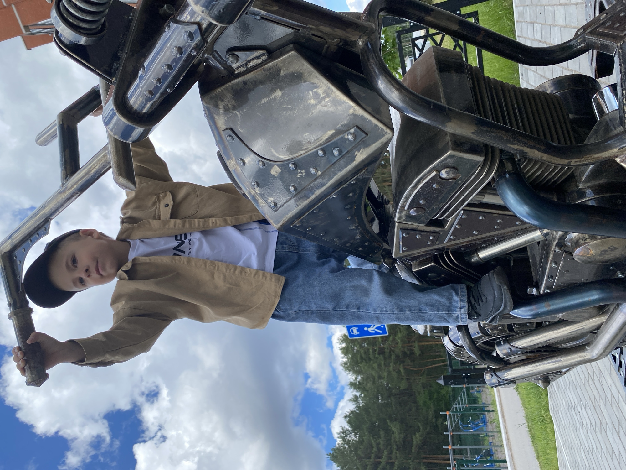 A low-angle, full-shot image shows a boy climbing on a metal sculpture in Chaikovsky, Russia. The boy, wearing a black cap, tan jacket, white shirt, and blue jeans, is holding onto the sculpture with both hands and has one foot resting on a pipe. The sculpture appears to be a stylized motorcycle made of welded metal. Behind the sculpture and boy are trees and a partly cloudy sky. A blue and white sign can be seen, next to a playground in the background. A stone path is visible on the right side of the frame, and a brick building can be seen behind the structure.