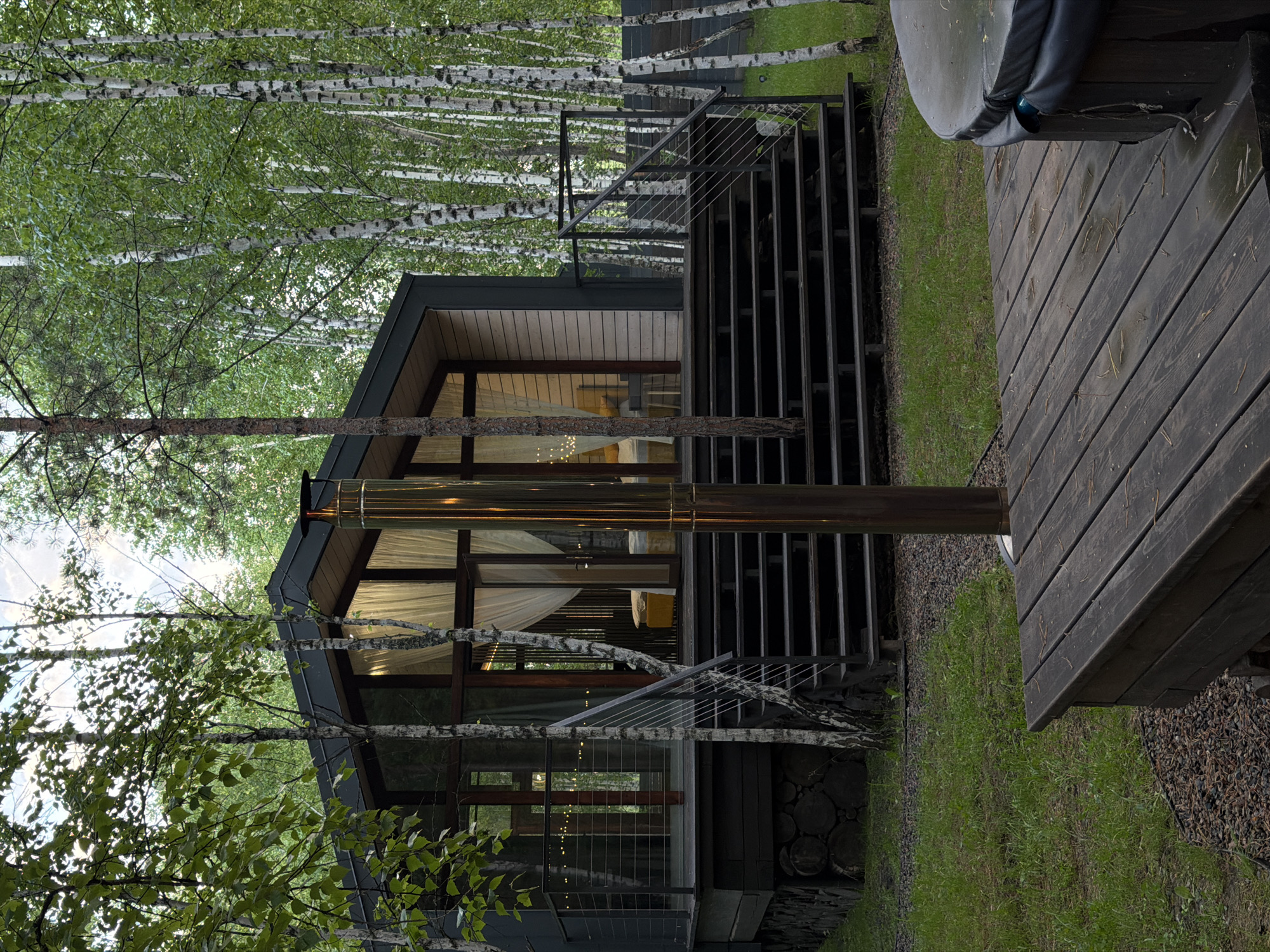 A modern, dark-sided, A-frame cabin with large glass windows and a light wood exterior sits nestled amongst a forest of birch trees. A wide set of dark wooden stairs leads up to a deck surrounding the cabin. A tall, polished metal chimney pipe stands prominently in the foreground. A dark wooden deck in the lower right corner partially shows a covered, round hot tub. The ground is covered with grass and gravel. The sky is overcast. The scene is located in Berezovka, Russia.