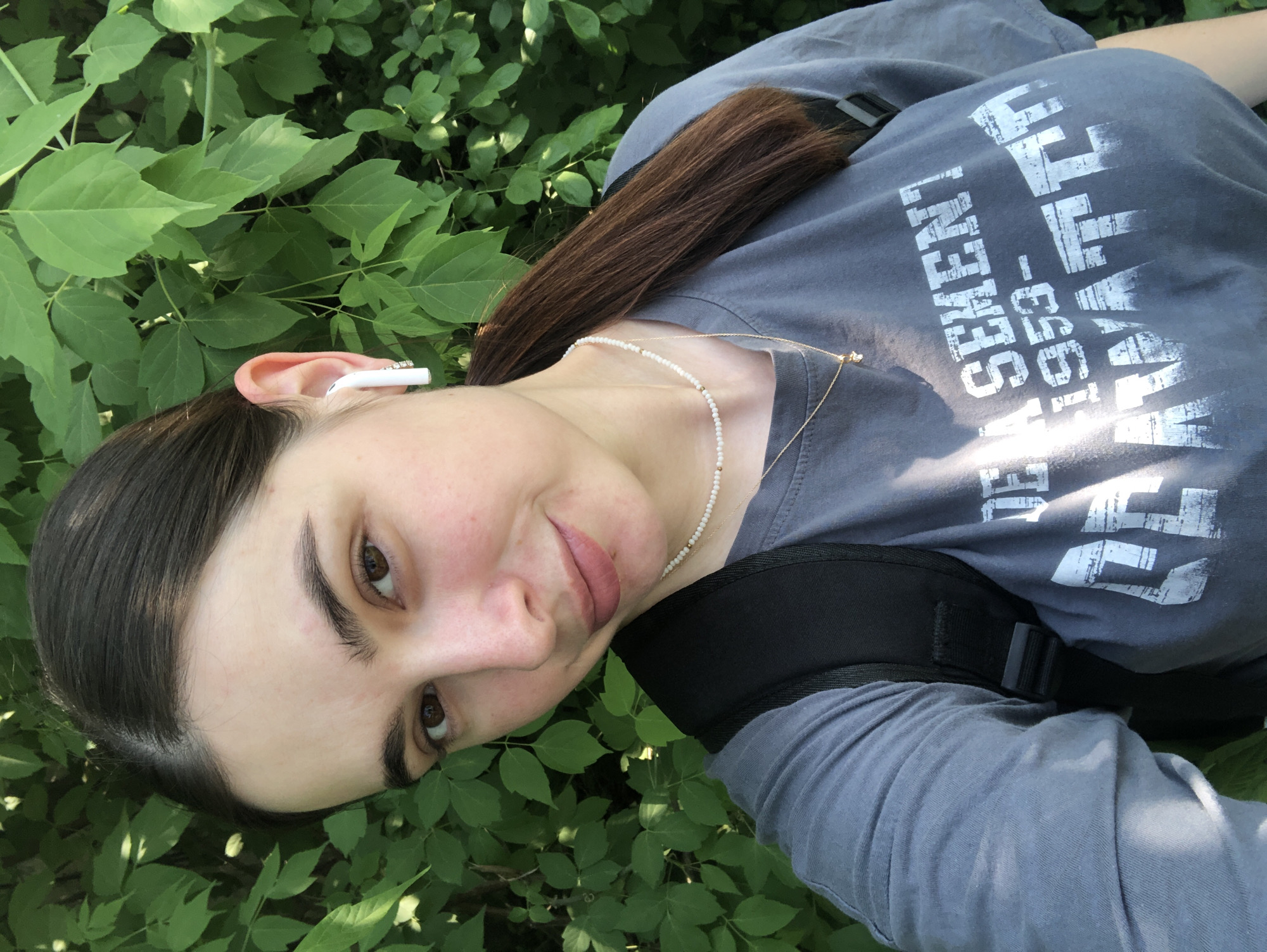 A young woman is visible in the image, positioned outdoors among lush green foliage, possibly in a park or garden. She has dark brown hair, fair skin, and brown eyes. She is wearing a gray t-shirt with white lettering, a pearl necklace, and a gold pendant necklace. An earbud is visible in her left ear. A backpack strap is visible on her shoulder. The location cannot be determined as Chaikovsky, Russia based on the visual information.