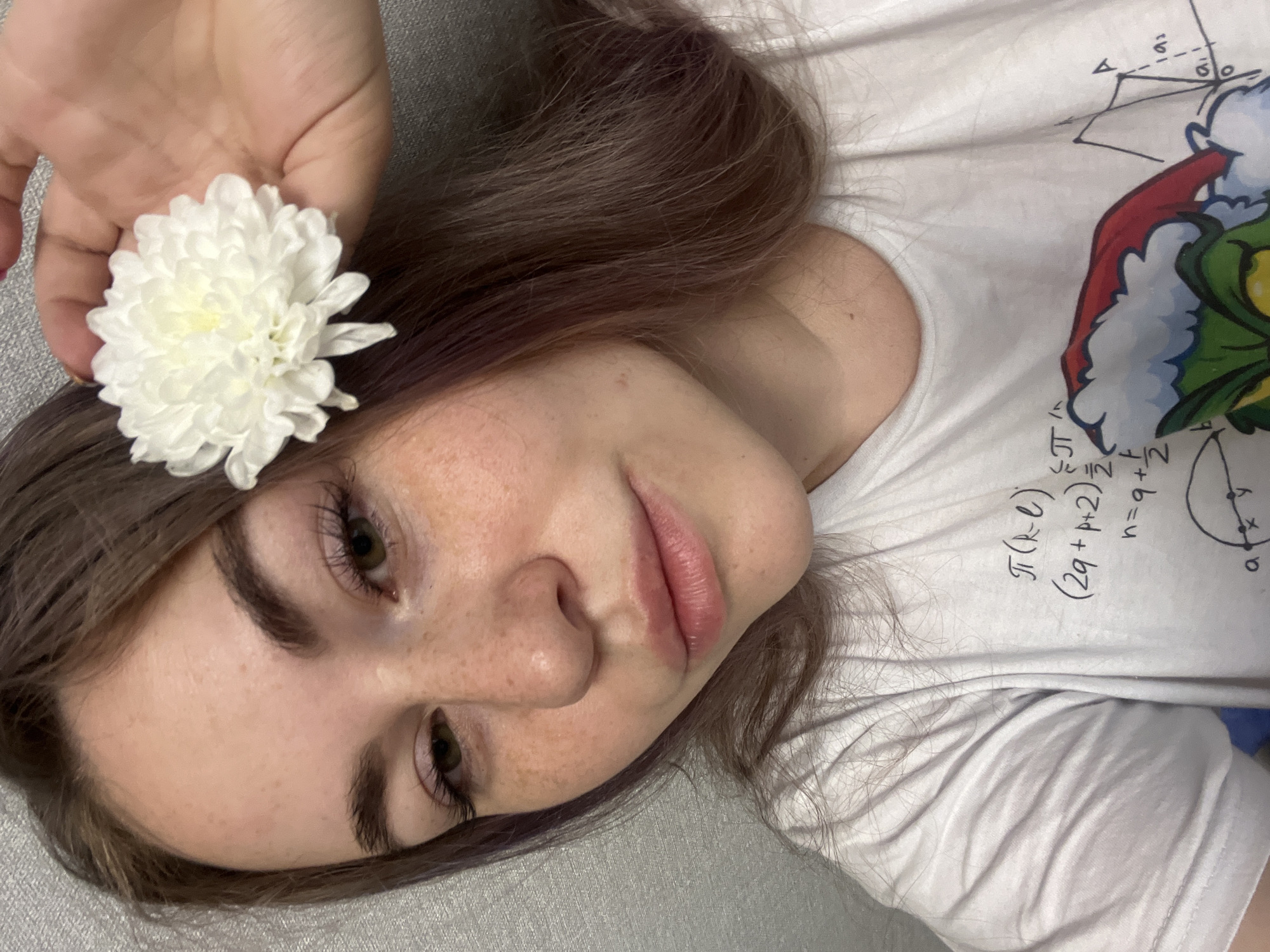 A young woman with light skin, brown hair, and freckles lies on a grey surface, possibly a couch or bed. A hand holds a white flower with many petals near her head, positioned above her forehead. She is wearing a white t-shirt featuring a cartoon illustration of the Grinch and mathematical equations. The equations include symbols such as π and variables such as 'n', 'q', 'p', 'k', and 'e'. No information indicates that this image was taken in the city of Chaikovsky, Russia.